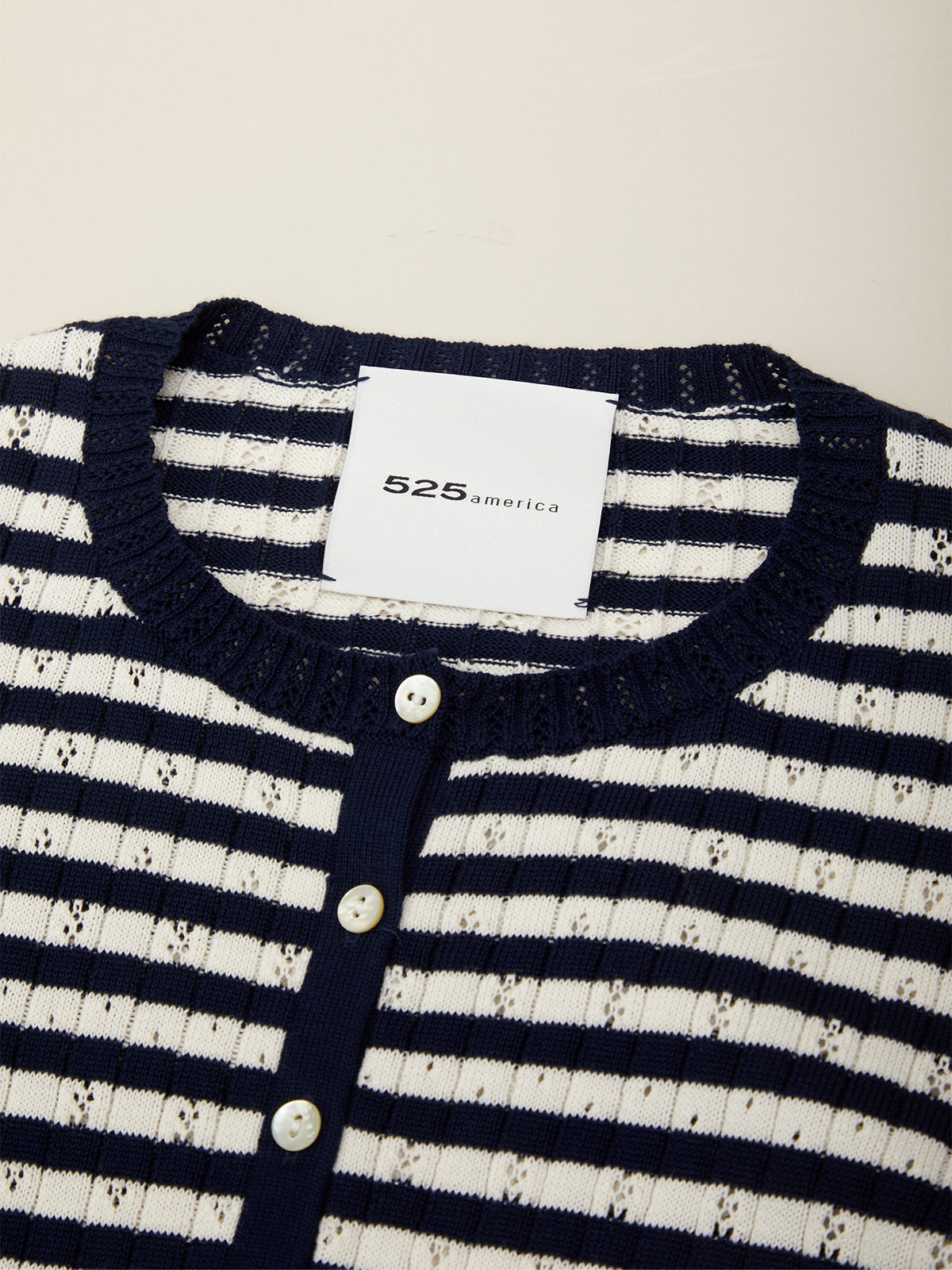 Close-up of a striped knit cardigan sweater with navy blue and white horizontal stripes, a navy ribbed crew neck, and a three-button placket. A white neck label reads 525america.