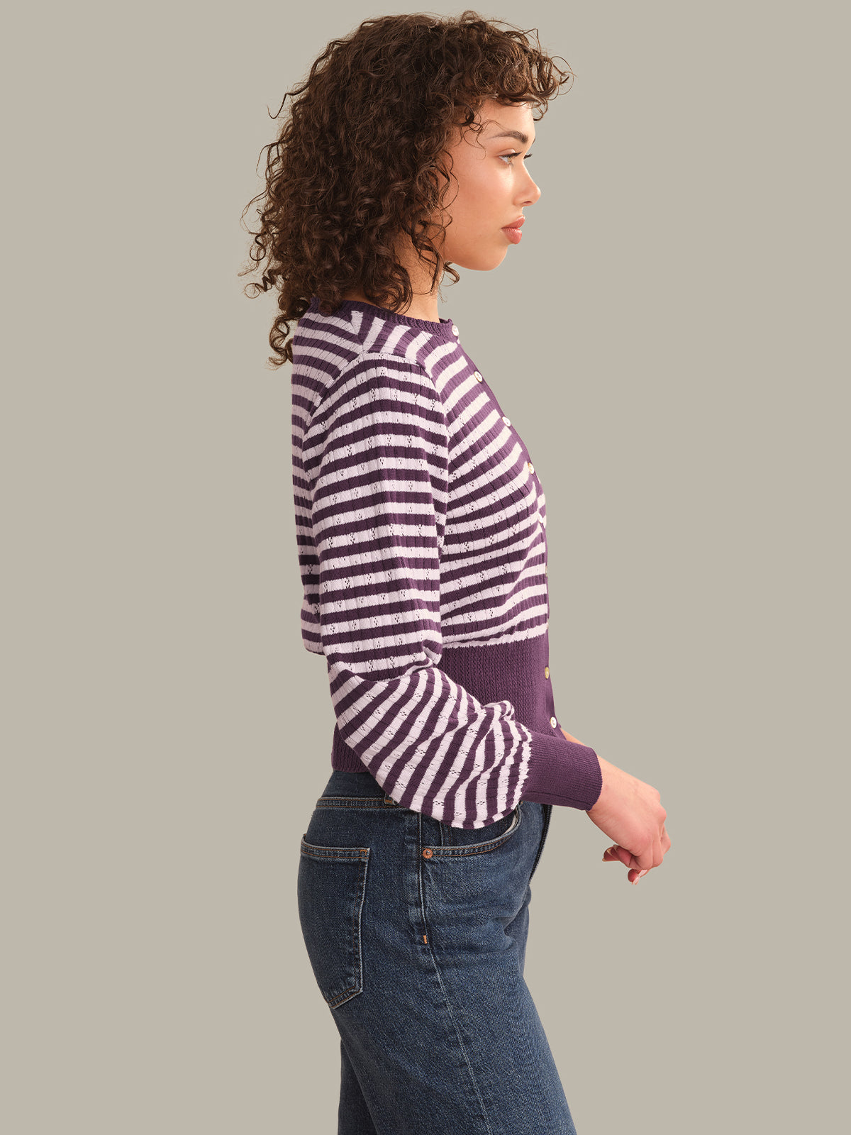 A woman shown in side profile wearing a purple and white horizontally striped long-sleeve knit cardigan with a solid purple waistband, paired with blue jeans, against a solid gray studio background.
