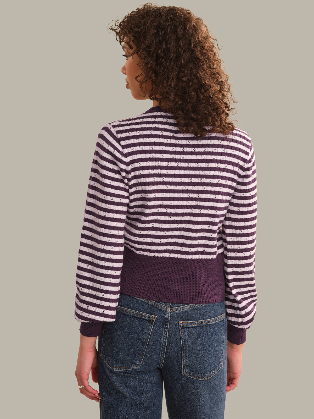A woman shown from the back wearing a cropped, long-sleeve knit cardigan with horizontal dark purple and off-white stripes, paired with blue jeans, against a solid light gray background.