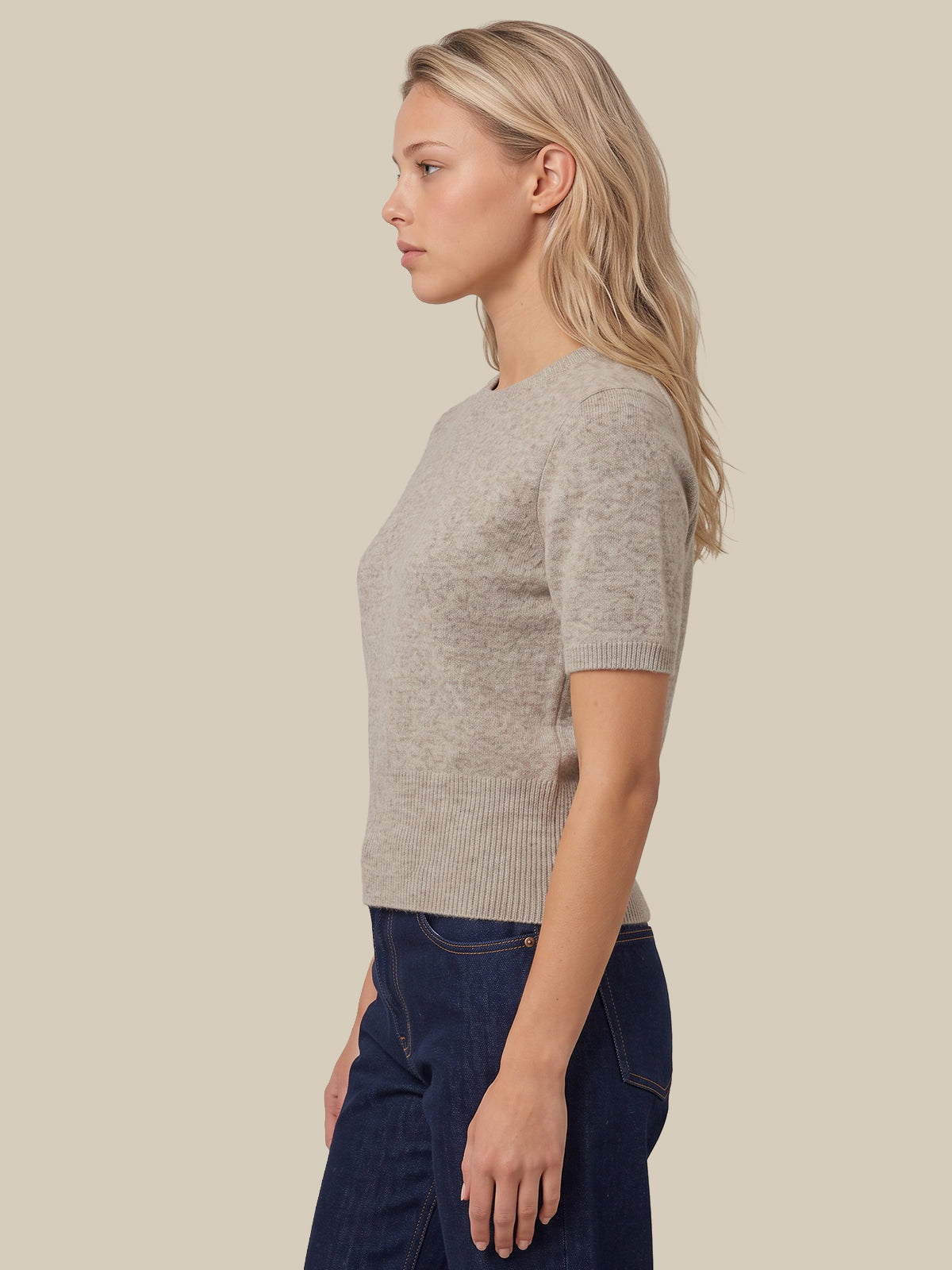 A woman in profile wearing a light beige short-sleeve knit top and dark blue jeans, photographed against a solid light beige background.