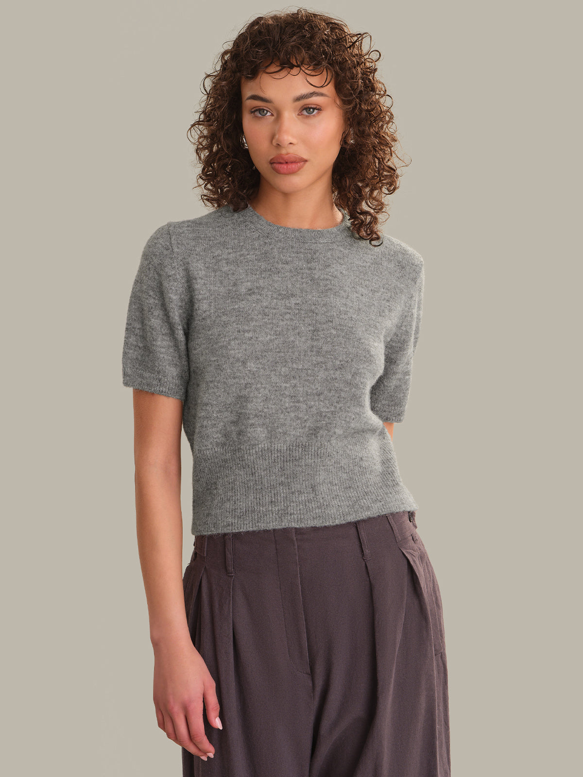 A model with curly brown hair wearing a short-sleeve gray knit top and dark brown pleated trousers, standing against a solid light gray background.