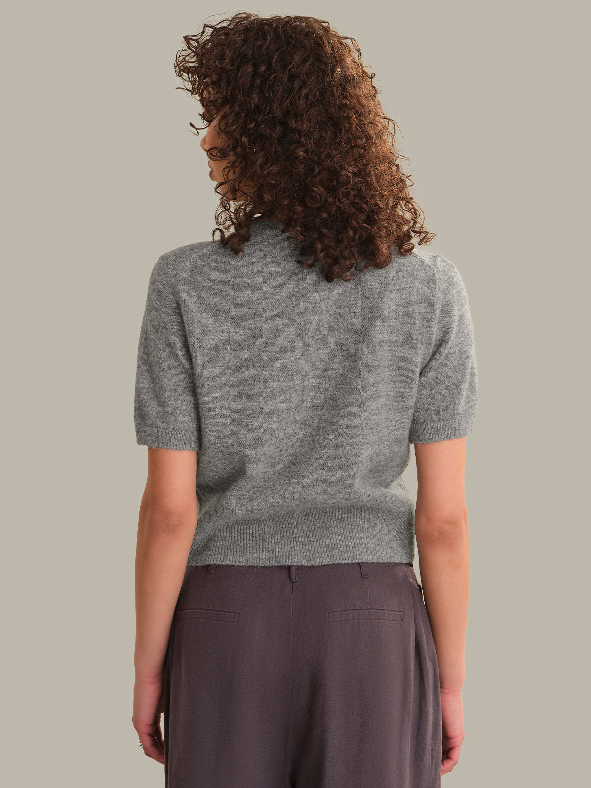 A woman with curly brown hair is shown from behind wearing a short-sleeve gray knit top and dark brown trousers, set against a solid light gray background.