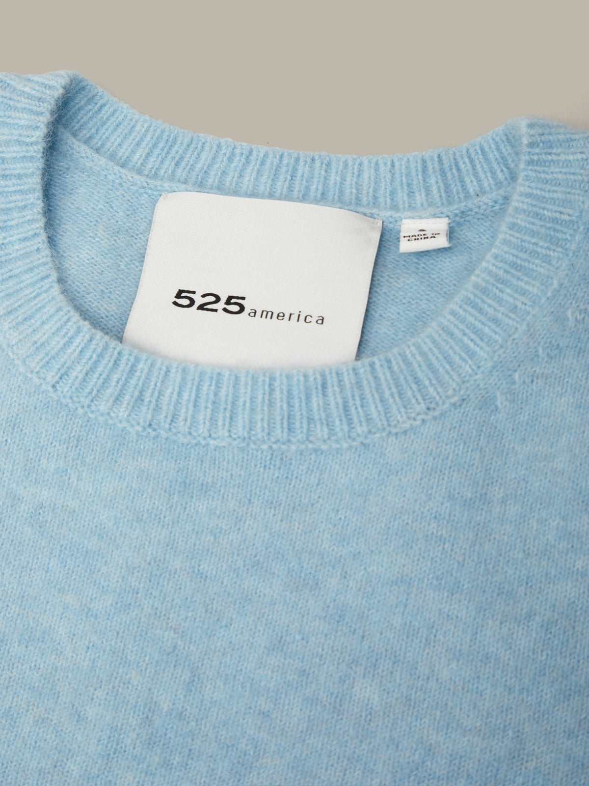 Close-up of a light blue knit sweater with a white neck label reading 525america, shown against a plain warm gray background.