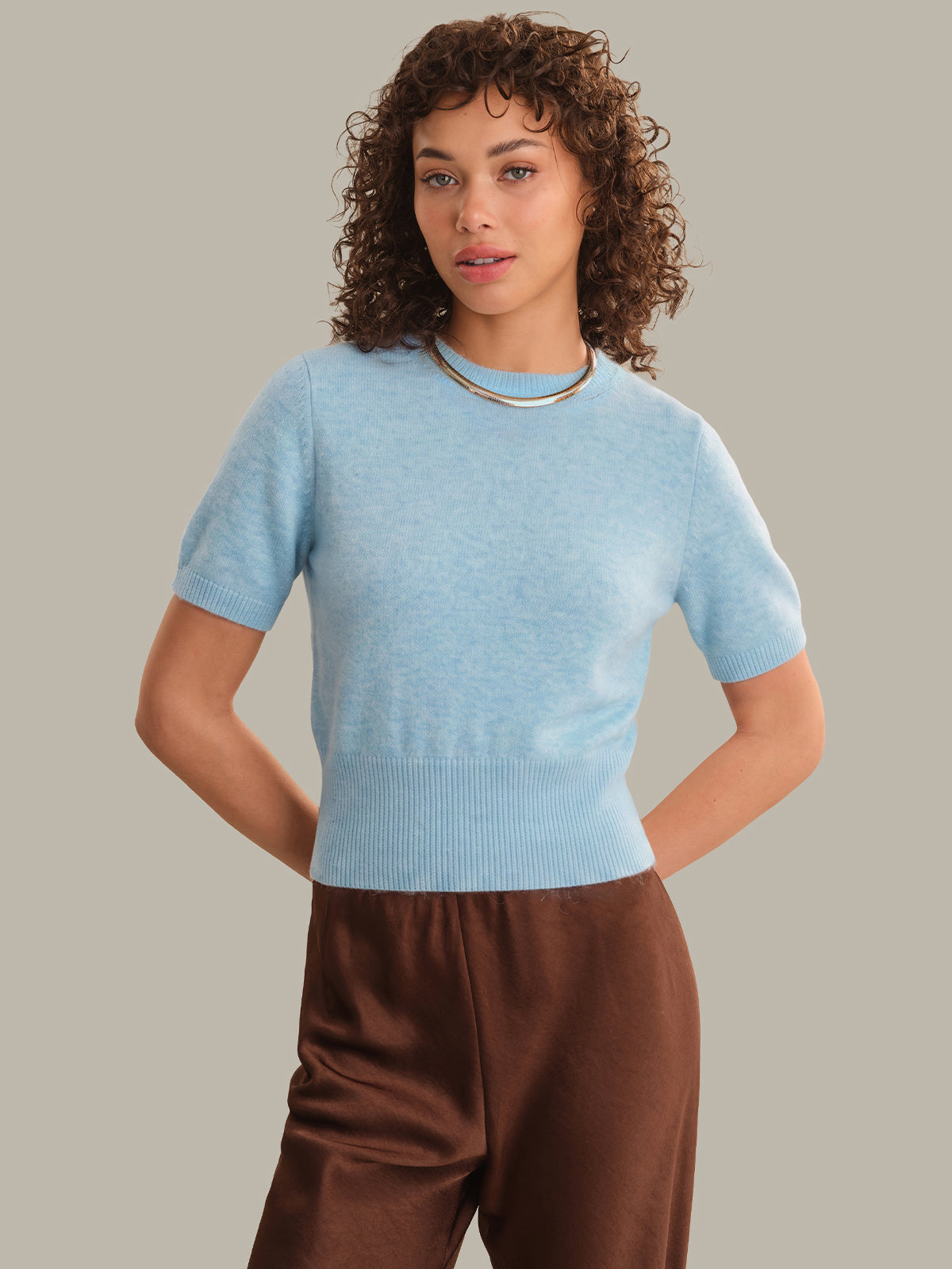 A woman with curly brown hair wearing a light blue short-sleeve knit top and brown pants, standing against a solid taupe background.