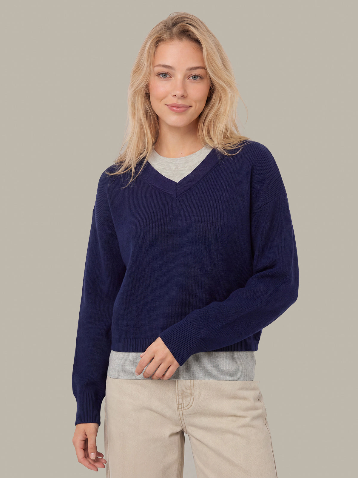 A blonde woman wearing a navy blue V-neck knit sweater over a light gray crewneck shirt, paired with beige pants, against a solid light gray background.