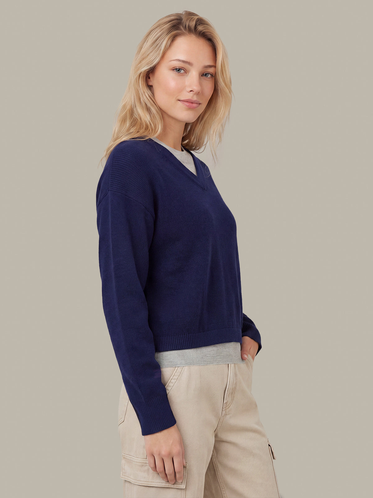 A woman wearing a navy blue V-neck knit sweater over a light gray undershirt, paired with beige cargo pants, standing against a solid taupe background.