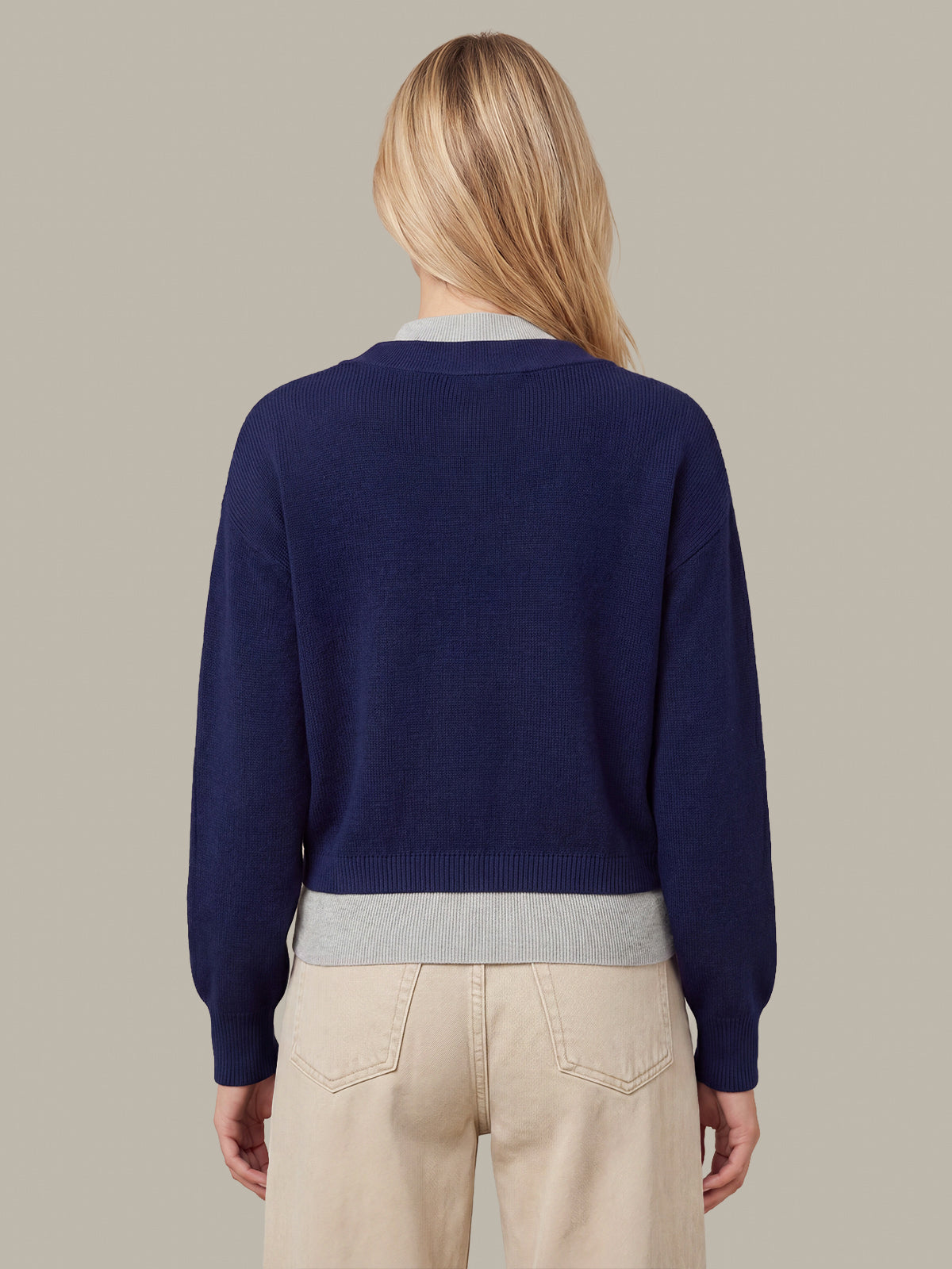 A woman viewed from the back wearing a navy blue knit sweater with light gray ribbed trim at the collar and hem, paired with beige pants, against a solid taupe background.