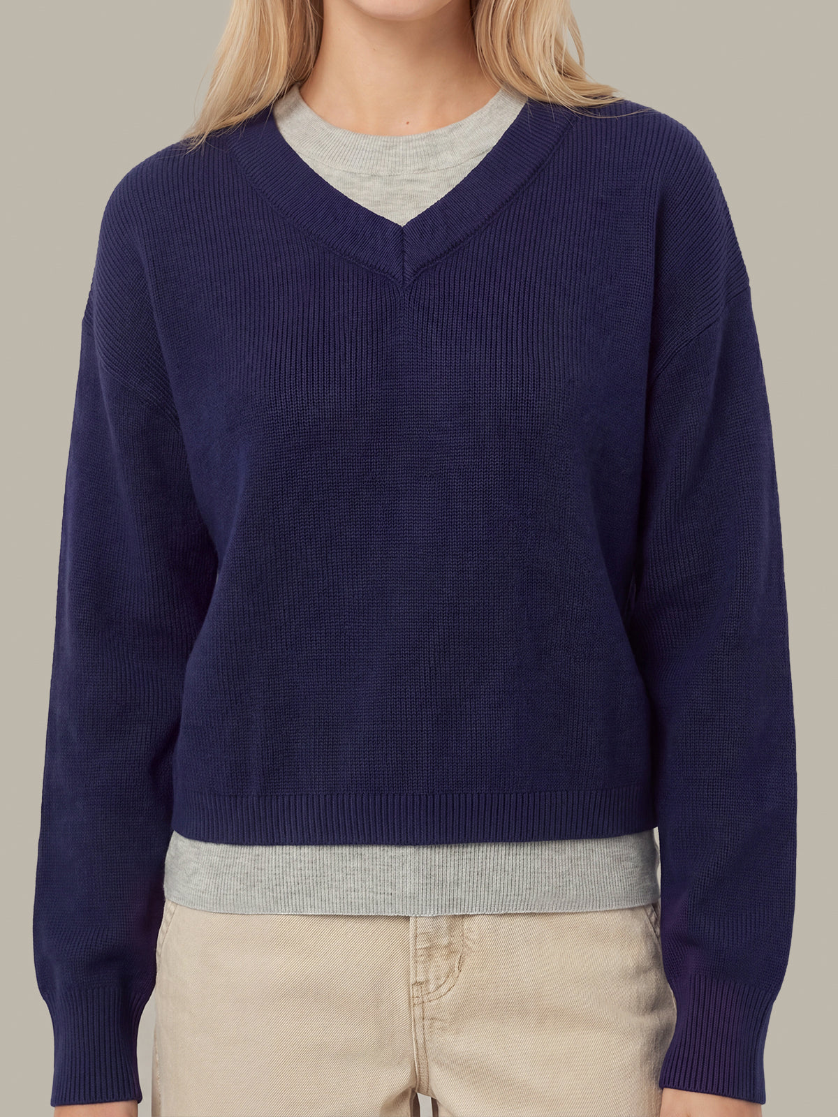 A woman wearing a navy blue ribbed V-neck sweater layered over a light gray crewneck shirt, paired with beige pants, against a solid light gray background.