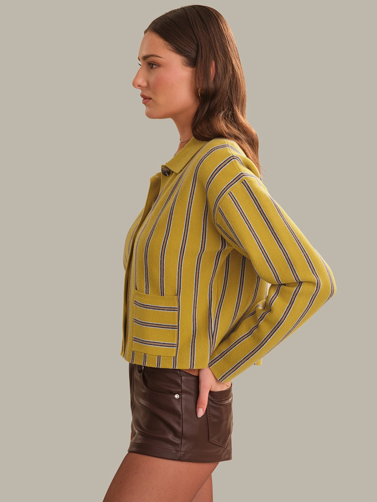 A woman shown in side profile wearing a cropped yellow striped cardigan with dark vertical stripes and brown leather shorts, against a solid light gray studio background.