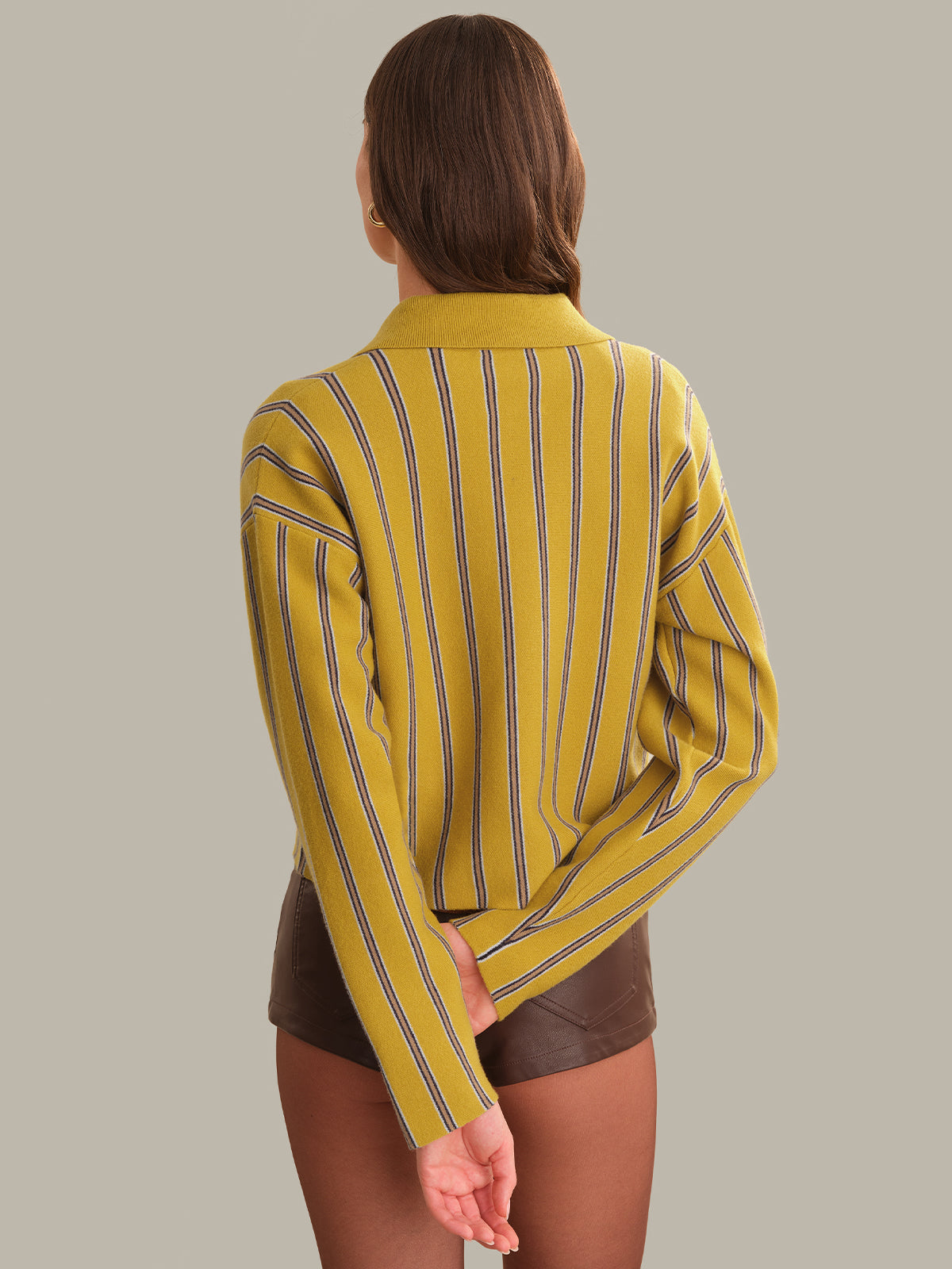 A woman shown from the back wearing a mustard yellow knit cardigan with thin vertical dark stripes and brown shorts, set against a solid light gray background.
