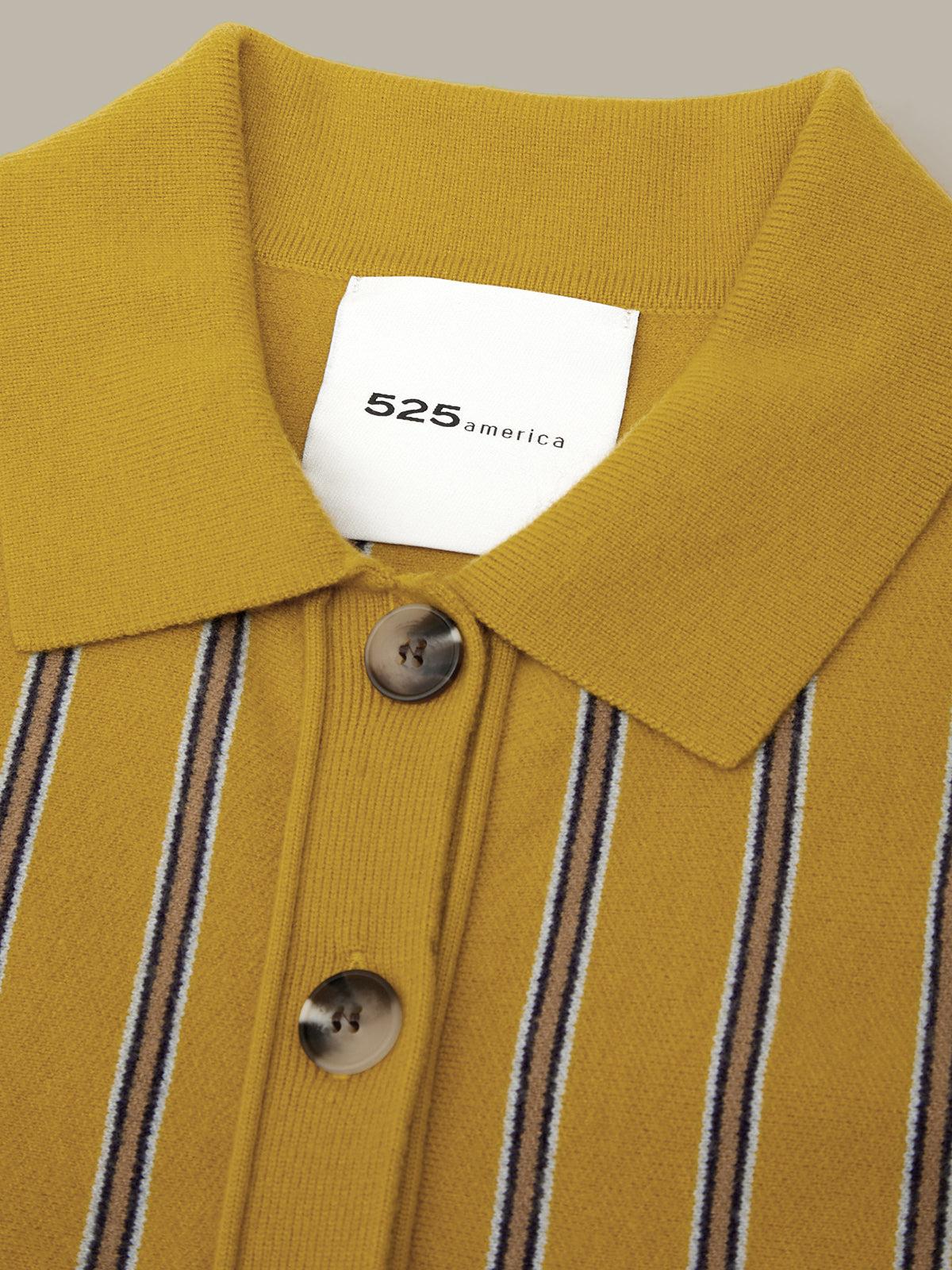 Close-up of a mustard yellow knit polo shirt with vertical brown and white stripes, featuring a white collar and a white neck label with the brand name '525 america'.