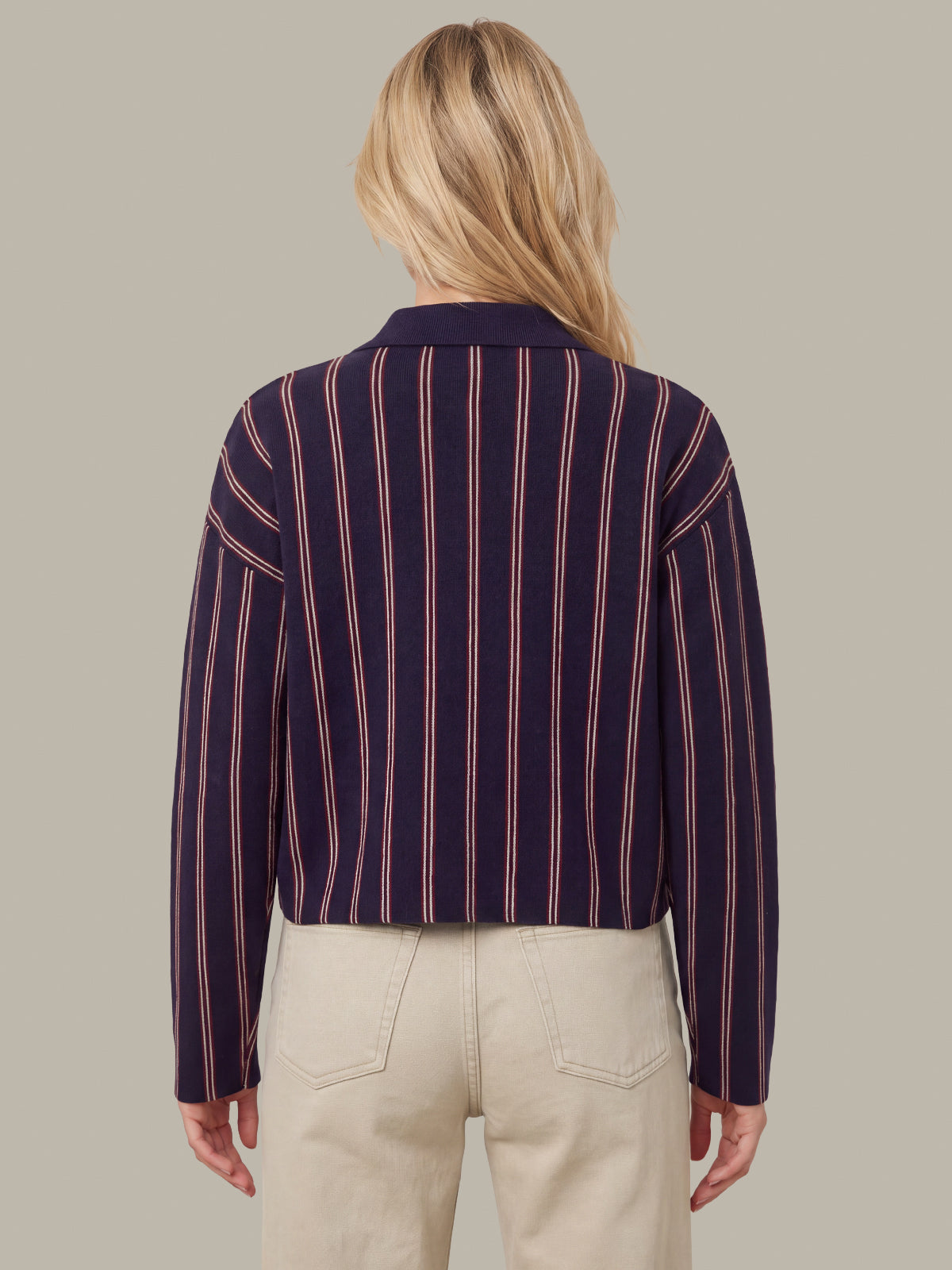 A woman viewed from the back wearing a cropped long-sleeve collared top with vertical navy, burgundy, and beige stripes, paired with light beige pants, against a solid taupe background.