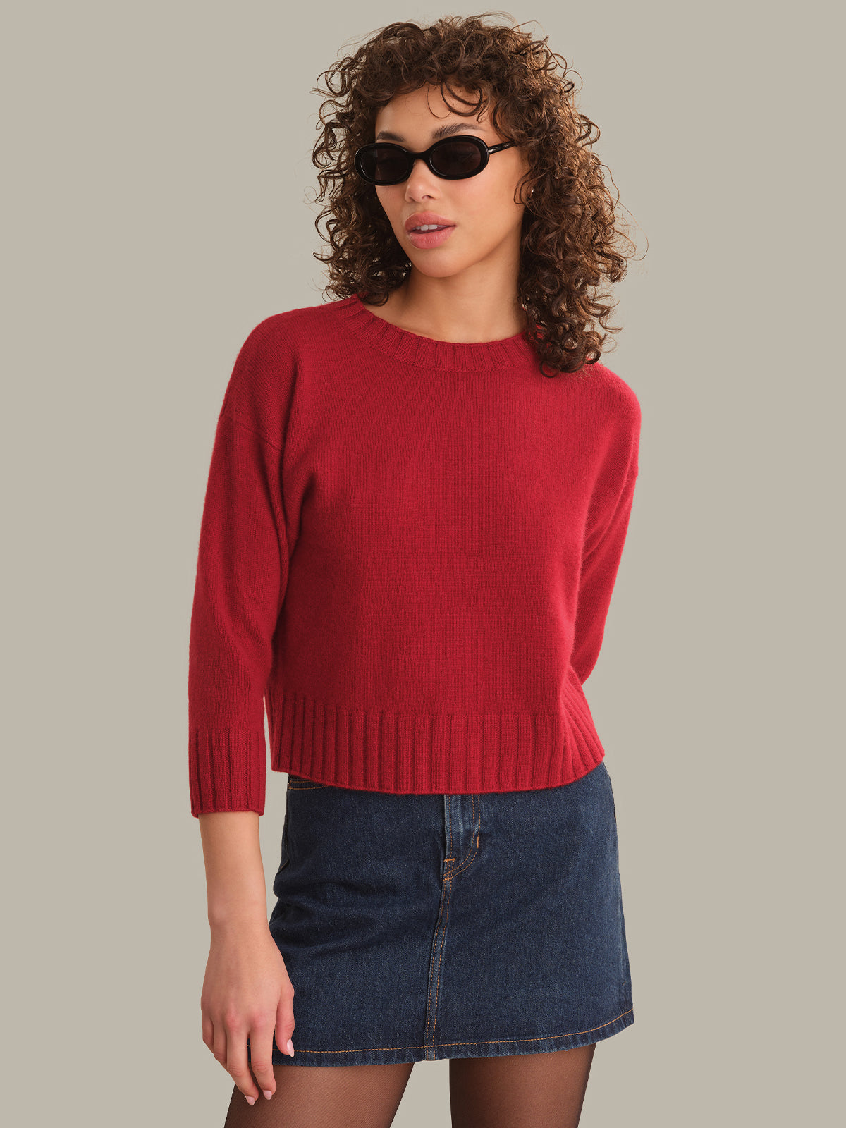 A woman with curly brown hair and black sunglasses wears a red knit sweater with three-quarter sleeves and a dark blue denim mini skirt, posed against a solid light gray background.