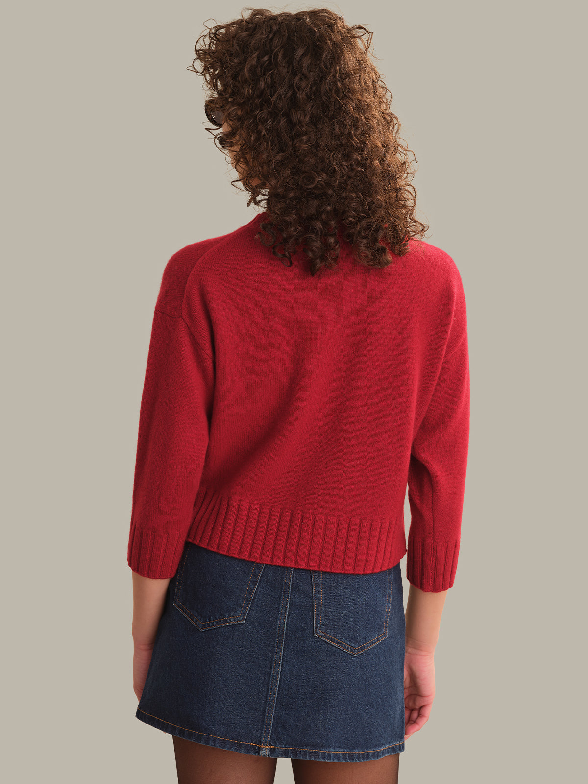 A woman shown from behind wearing a bright red knit sweater and a blue denim mini skirt against a solid light gray background.
