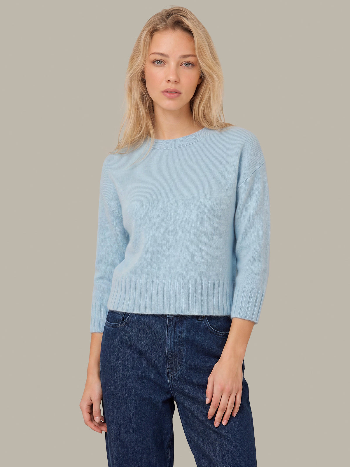 A blonde woman wearing a light blue knit sweater and dark blue jeans, standing against a solid taupe background.
