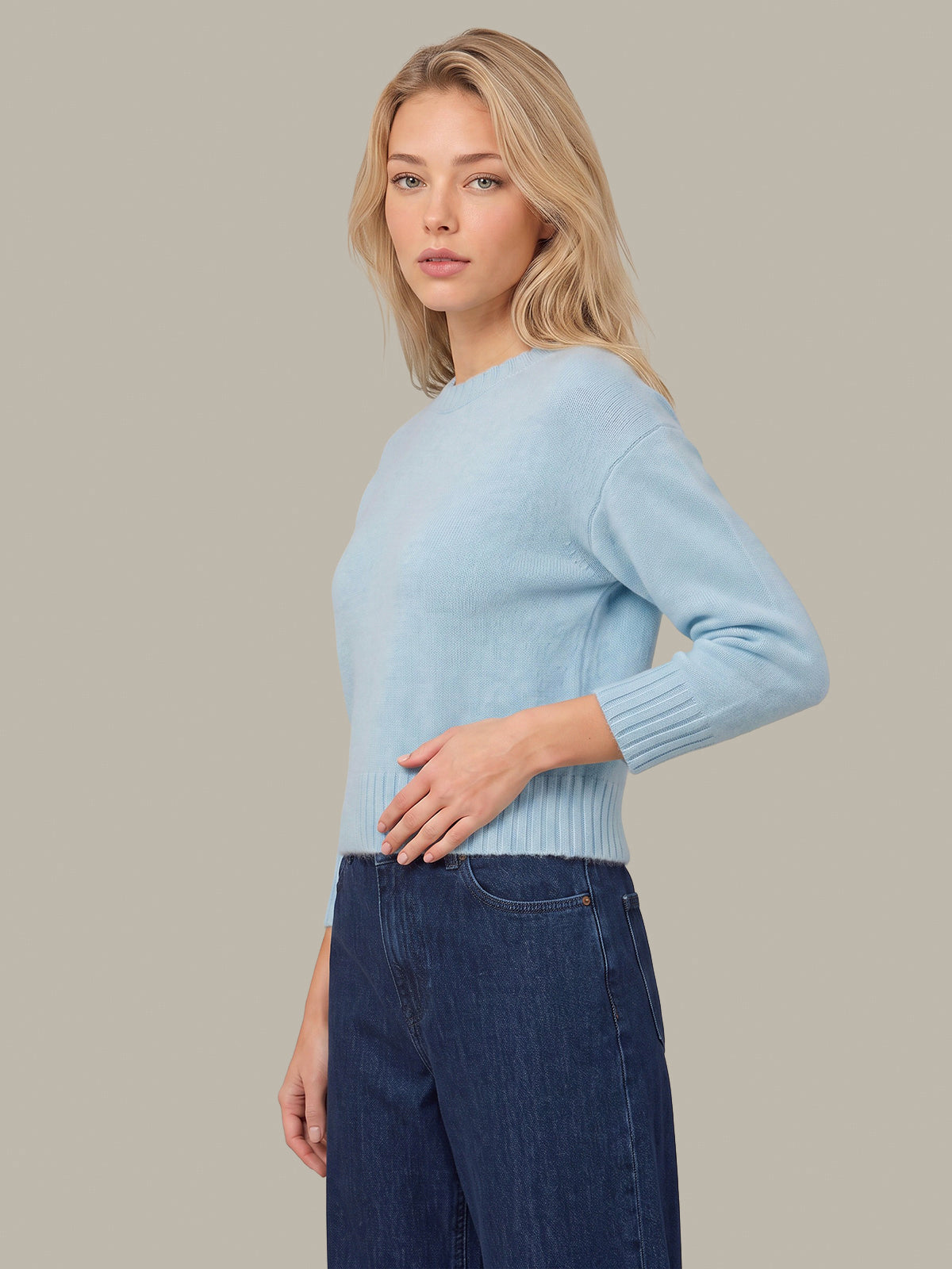 A blonde woman wearing a light blue knit sweater and dark blue jeans, shown in a side-profile studio shot against a solid beige background.