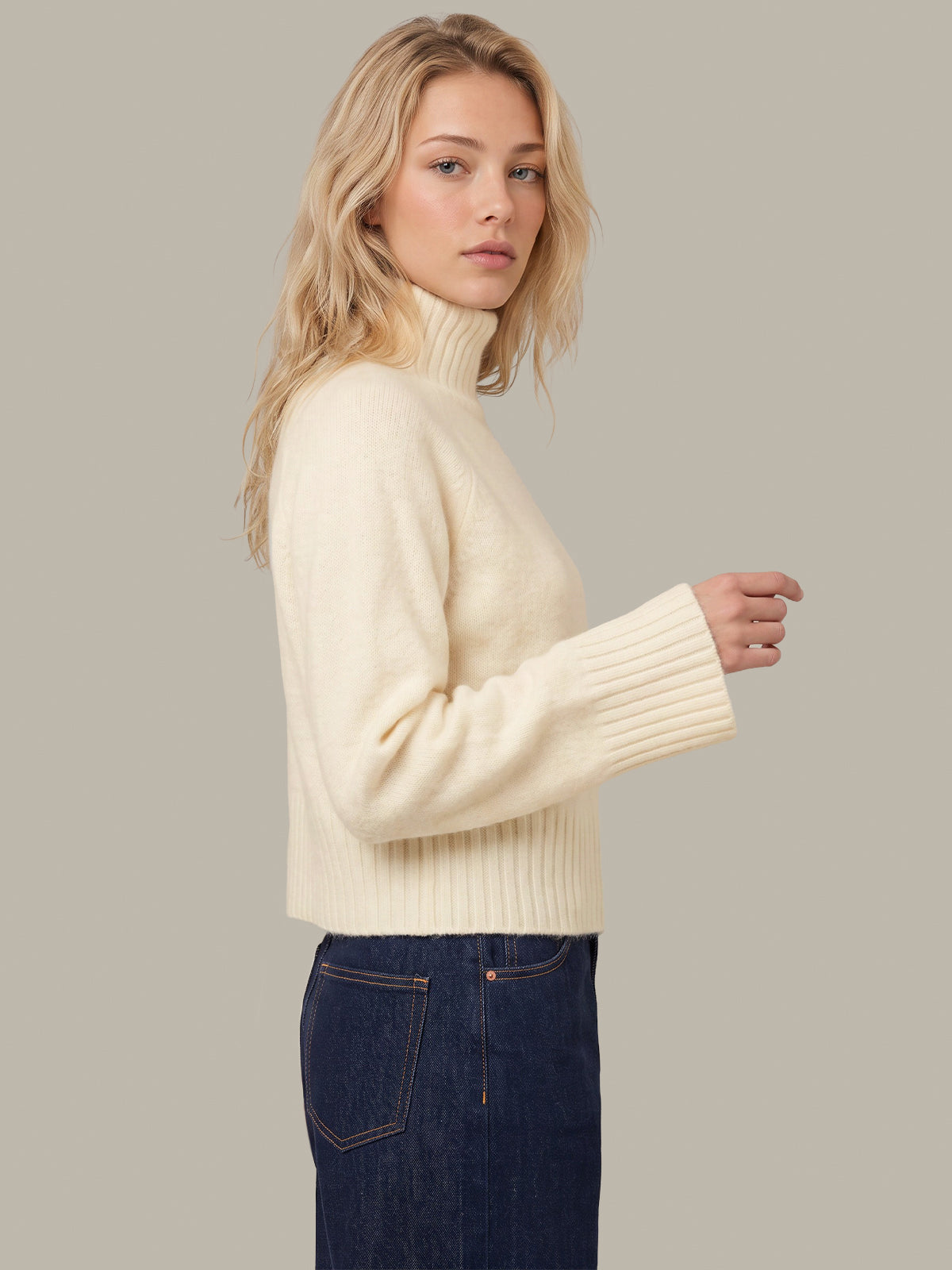 A woman wearing a cream ribbed turtleneck sweater and dark blue jeans, shown from a side profile against a solid light gray background.