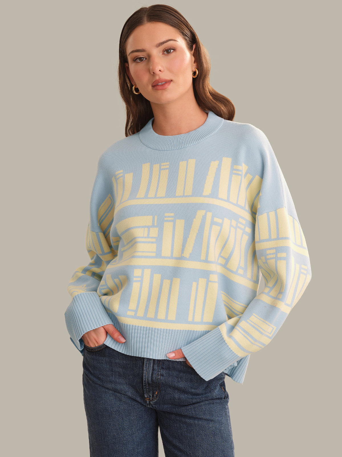 A woman wearing a light blue crewneck sweater with a pale yellow bookshelf pattern, paired with dark blue jeans, against a solid gray background.