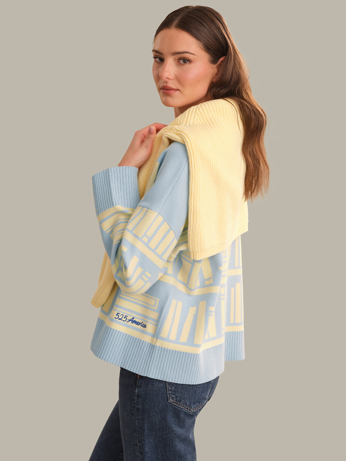 A woman wearing a light blue and pale yellow knit sweater with geometric patterns and a matching yellow knit wrap draped over her shoulders, paired with dark blue jeans, against a solid gray background.