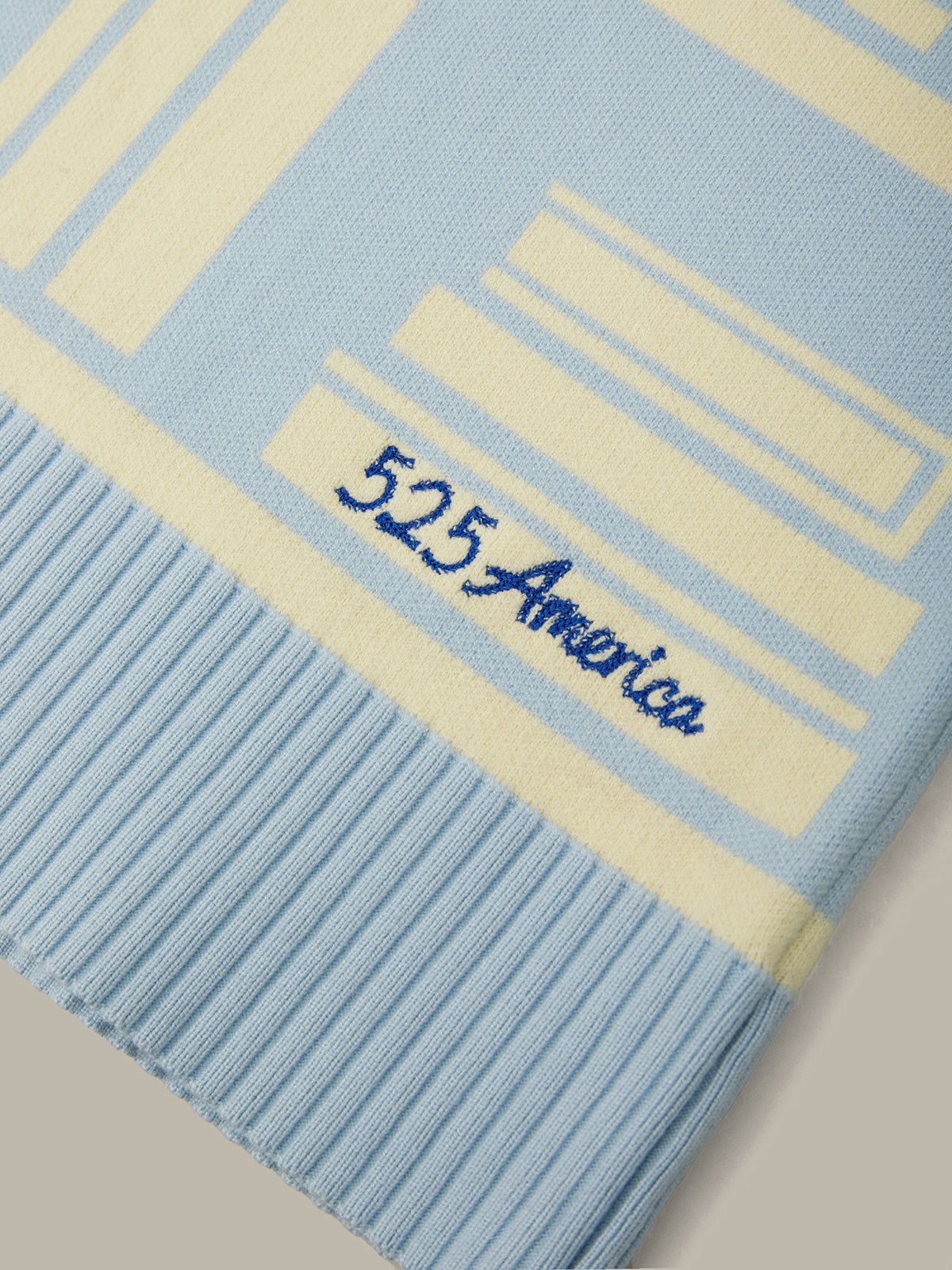 A close-up of a light blue and cream striped knit scarf with a ribbed border and the embroidered brand name '525 America' in blue script.