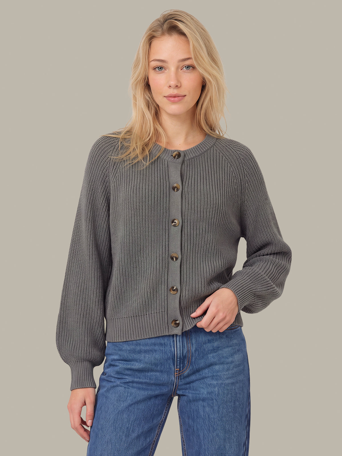 A blonde woman wearing a dark gray ribbed knit cardigan with brown buttons and blue jeans, standing against a solid taupe background.