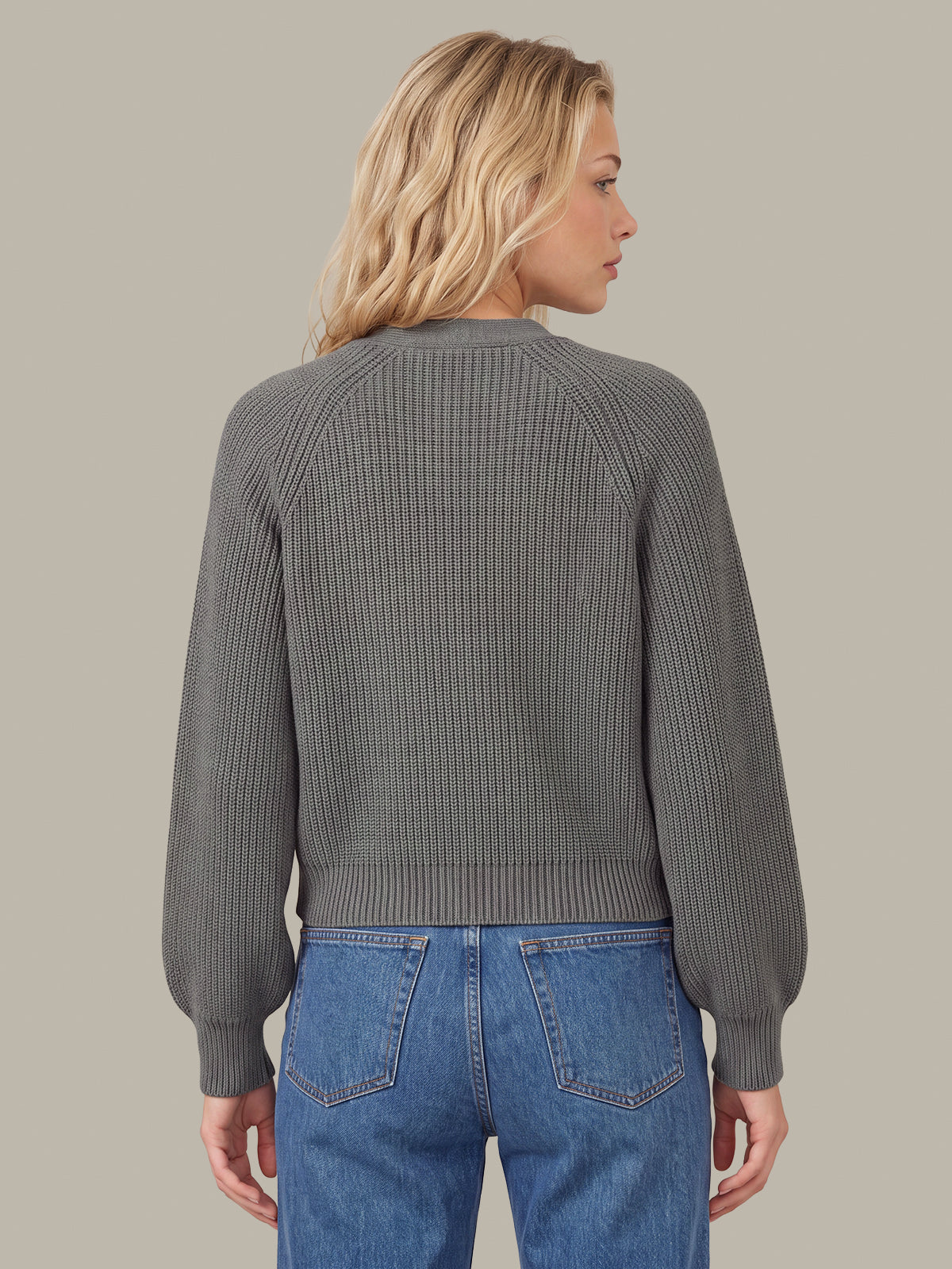 A woman shown from the back wearing a gray ribbed knit sweater and blue jeans against a solid taupe background.