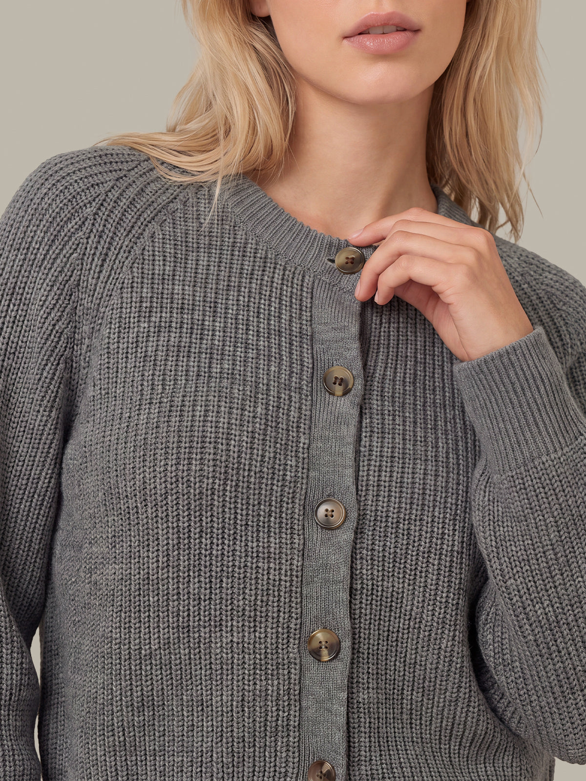 A close-up of a woman wearing a gray ribbed knit cardigan sweater with brown buttons, cropped from the mouth down against a plain beige background.