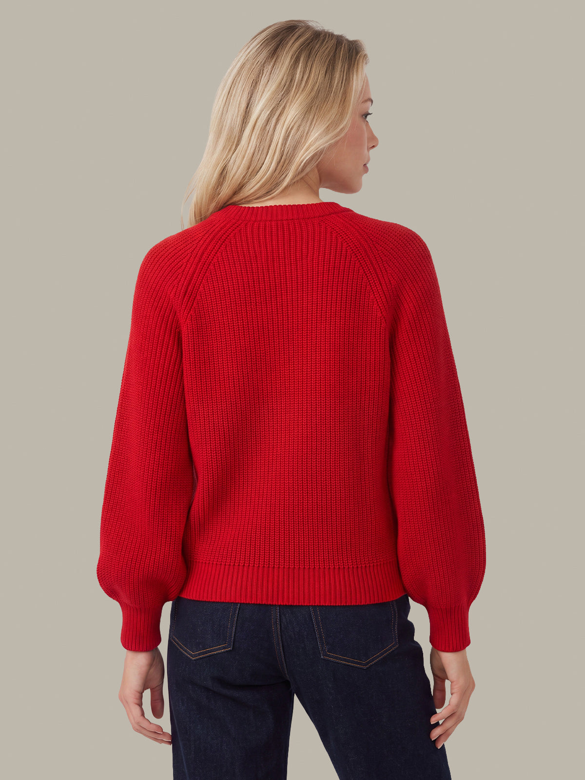 A woman shown from the back wearing a bright red ribbed knit sweater with balloon sleeves and dark blue jeans, set against a solid light gray background.