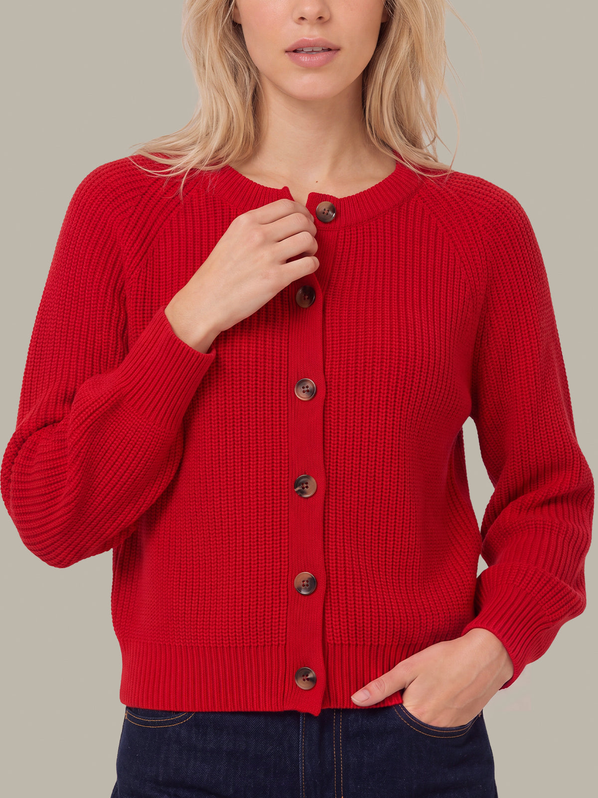 A woman wearing a bright red ribbed knit cardigan sweater with dark buttons, paired with dark blue jeans, against a solid light gray background.