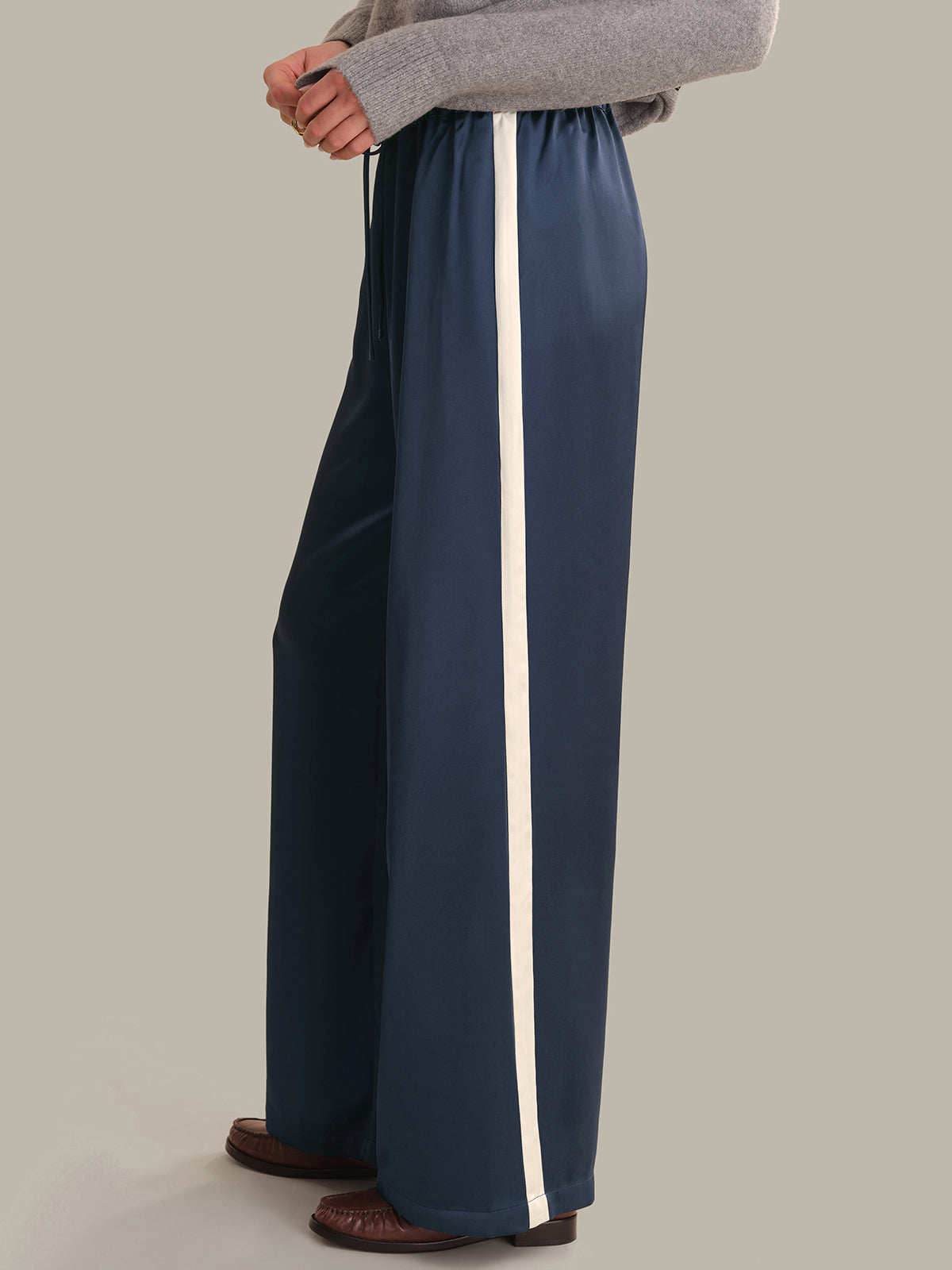A person wearing navy wide-leg pants with a vertical white stripe down the side, paired with a gray sweater and brown shoes, shown from the waist down against a plain light gray studio background.