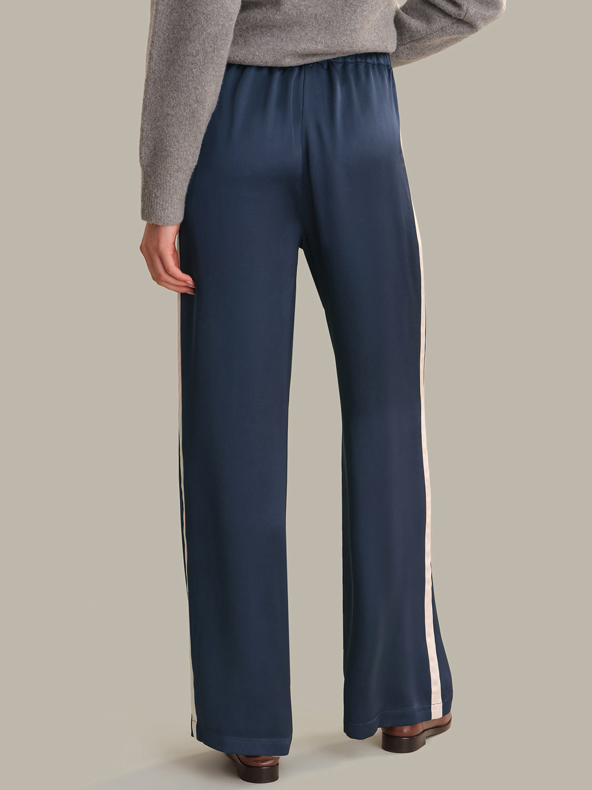 A person wearing navy wide-leg pants with white side stripes, paired with a gray sweater and brown shoes, shown from the back against a plain beige studio background.