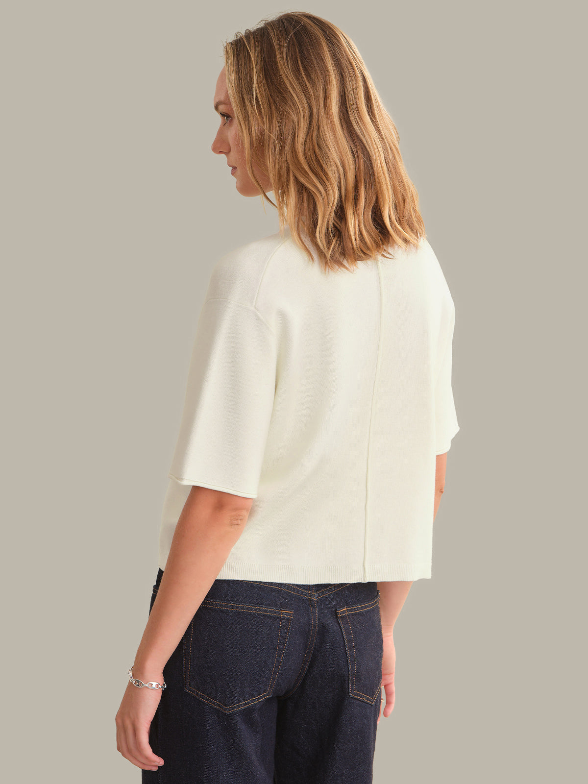 A woman shown from the back wearing a short-sleeve cream-colored knit top and dark blue jeans, set against a solid neutral gray background.