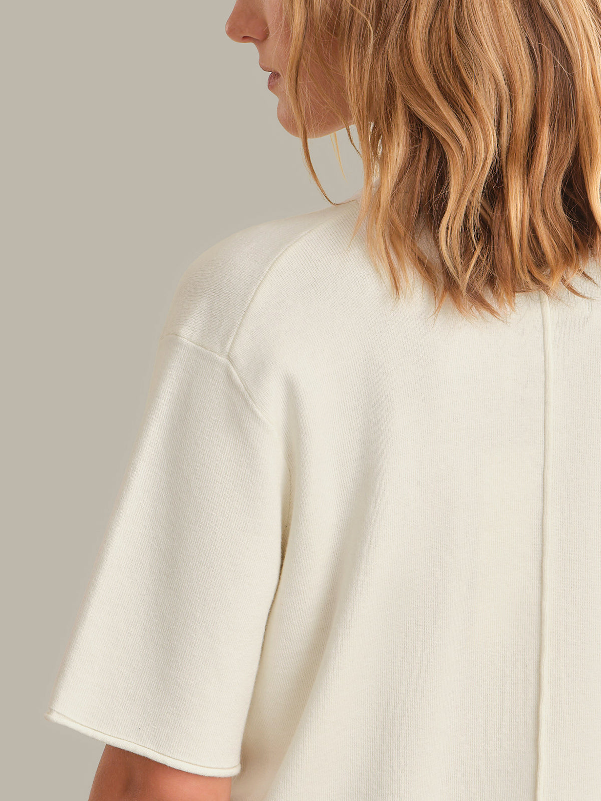 Close-up back view of a woman wearing a cream-colored short-sleeve knit top against a plain light gray background.
