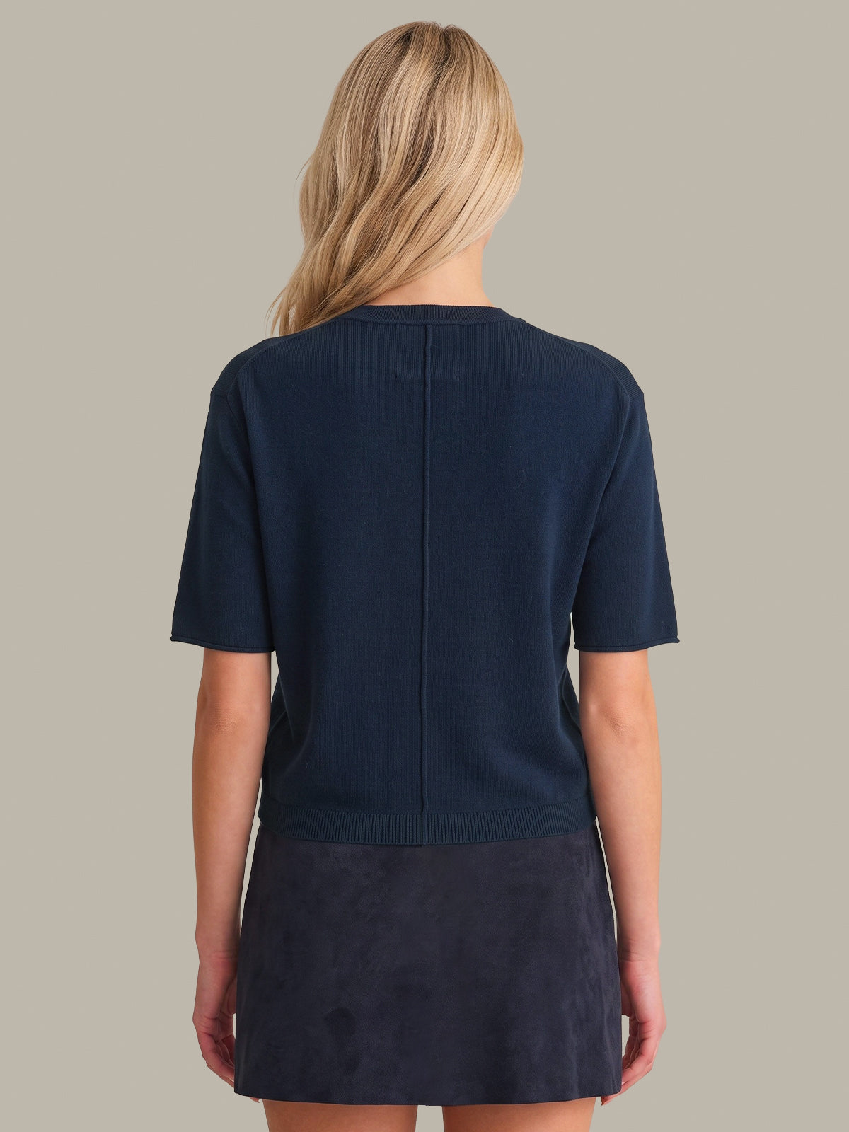 A woman viewed from the back wearing a dark navy short-sleeve knit top and matching dark skirt against a plain warm gray studio background.