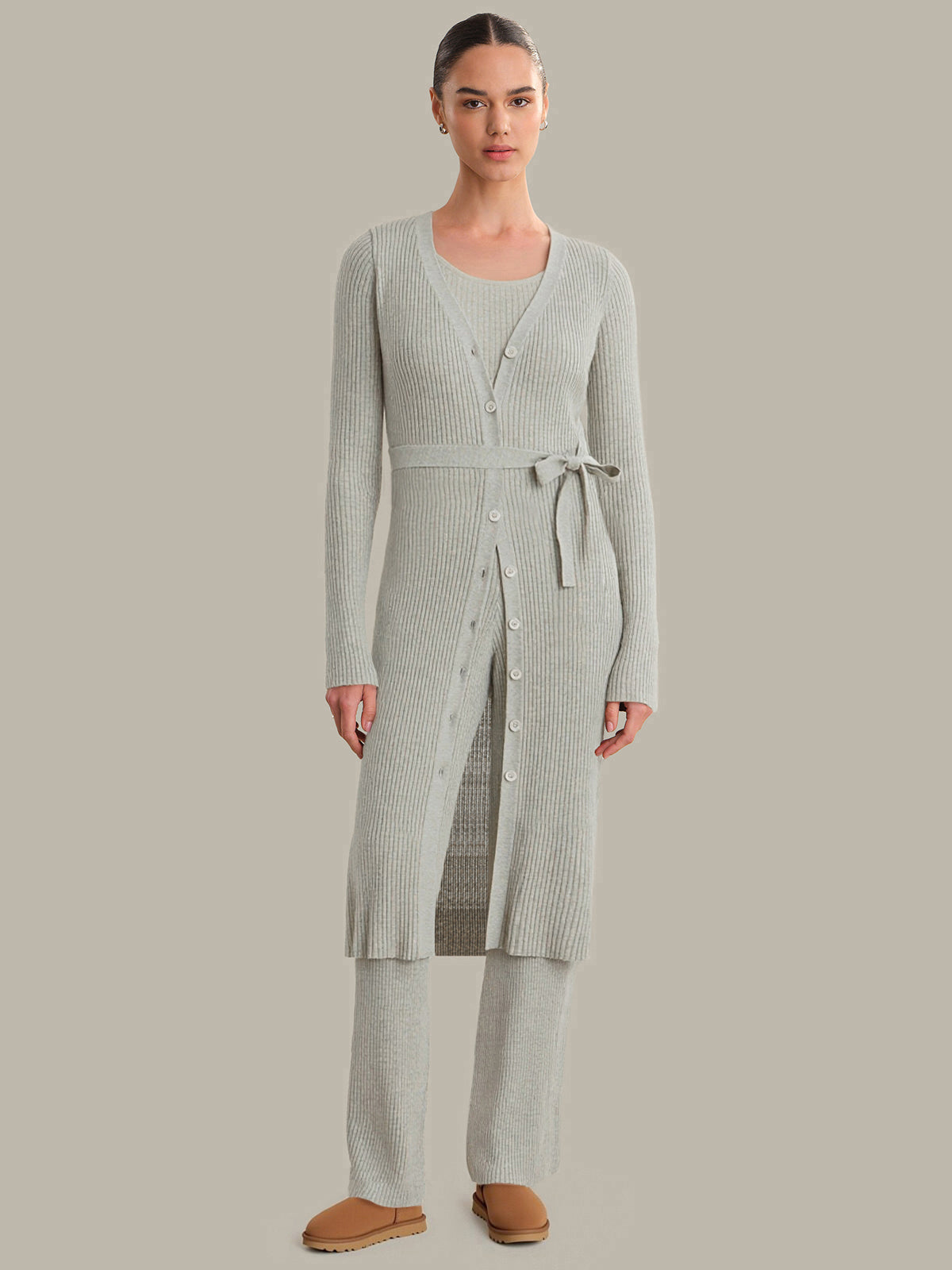Woman wearing a long, light gray cardigan over a matching dress with a belt on a plain background