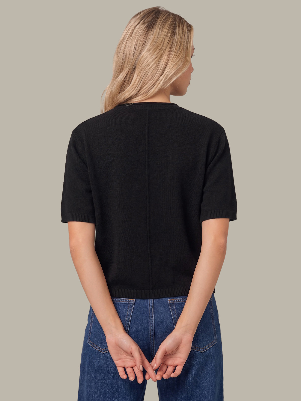 A woman viewed from the back wearing a black short-sleeve knit top and blue jeans against a plain warm gray studio background.
