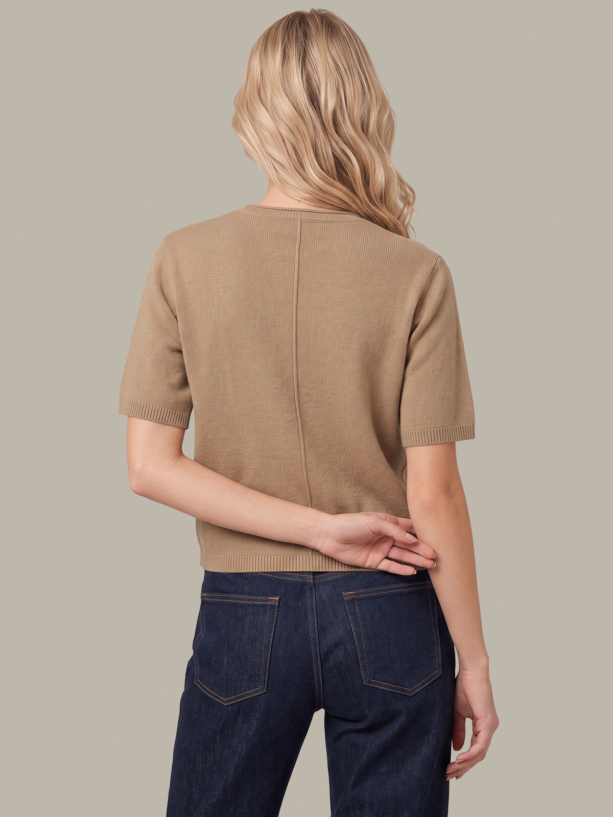 A woman shown from the back wearing a short-sleeve knit top in a warm tan/camel color, paired with dark blue jeans, against a solid light gray background.