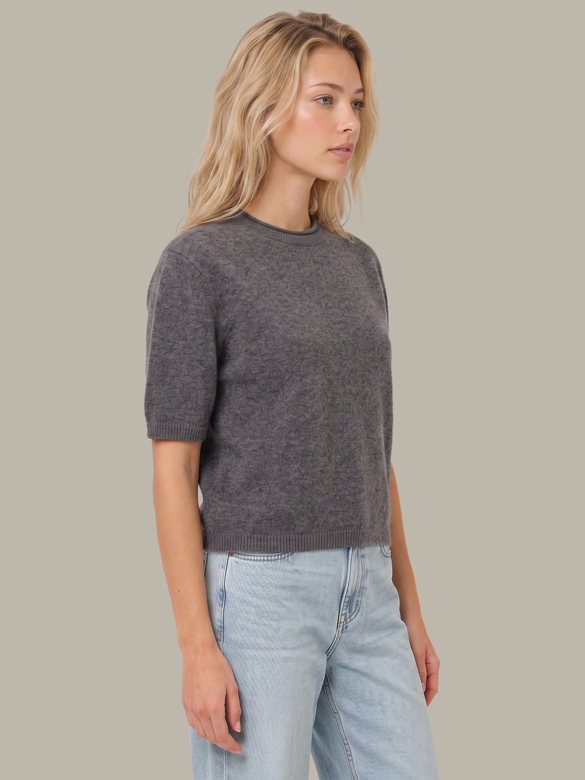 A woman wearing a dark gray short-sleeve knit sweater and light blue jeans, standing against a solid light gray background.