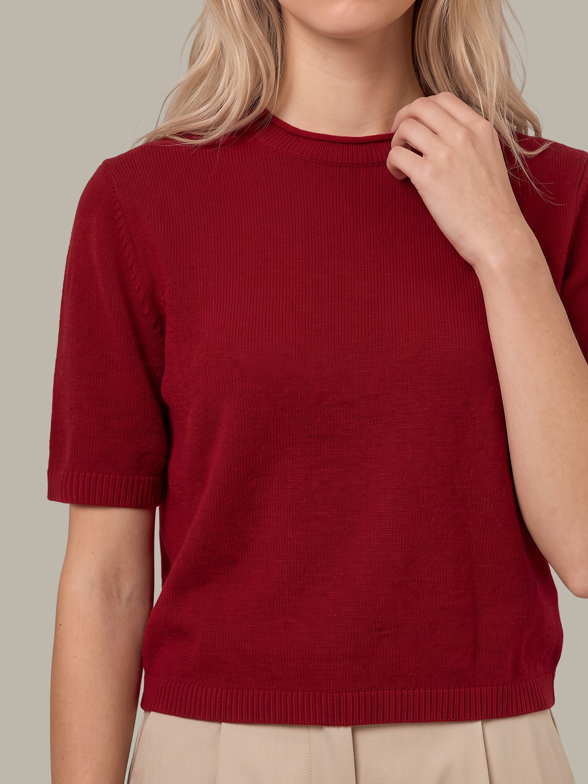 A close-up of a woman wearing a short-sleeve ribbed knit sweater in deep burgundy red, paired with beige trousers, against a solid light gray background.