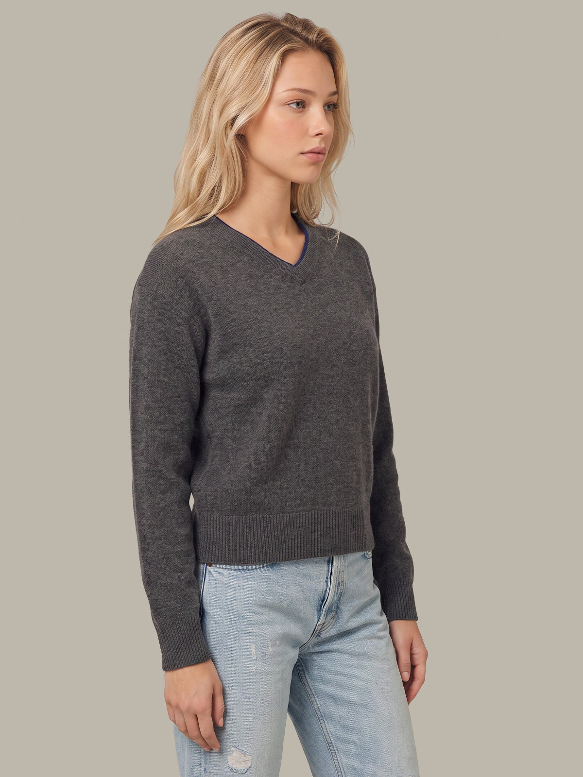A woman wearing a dark gray heathered V-neck knit sweater and light-wash blue jeans, standing against a solid light gray studio background.