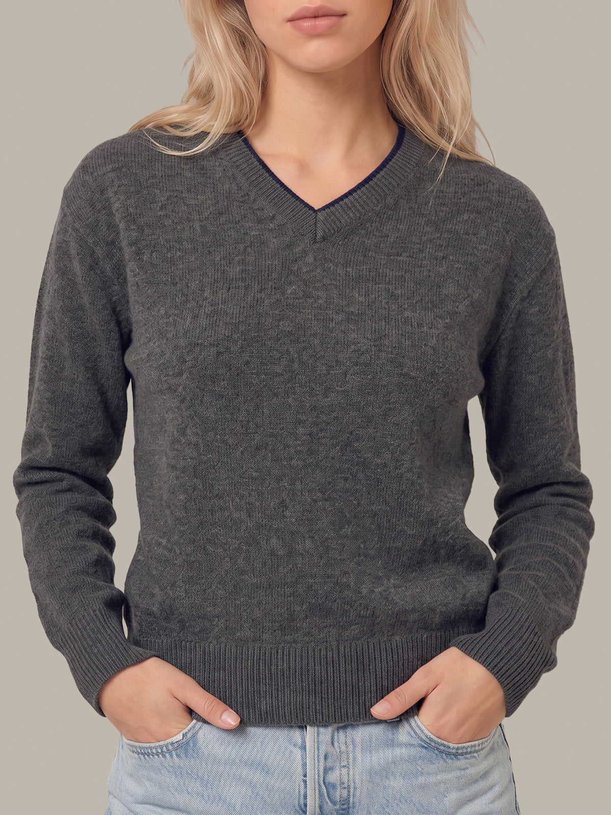 A woman wearing a dark gray heathered v-neck knit sweater with ribbed cuffs and hem, paired with light blue jeans, against a solid neutral background.