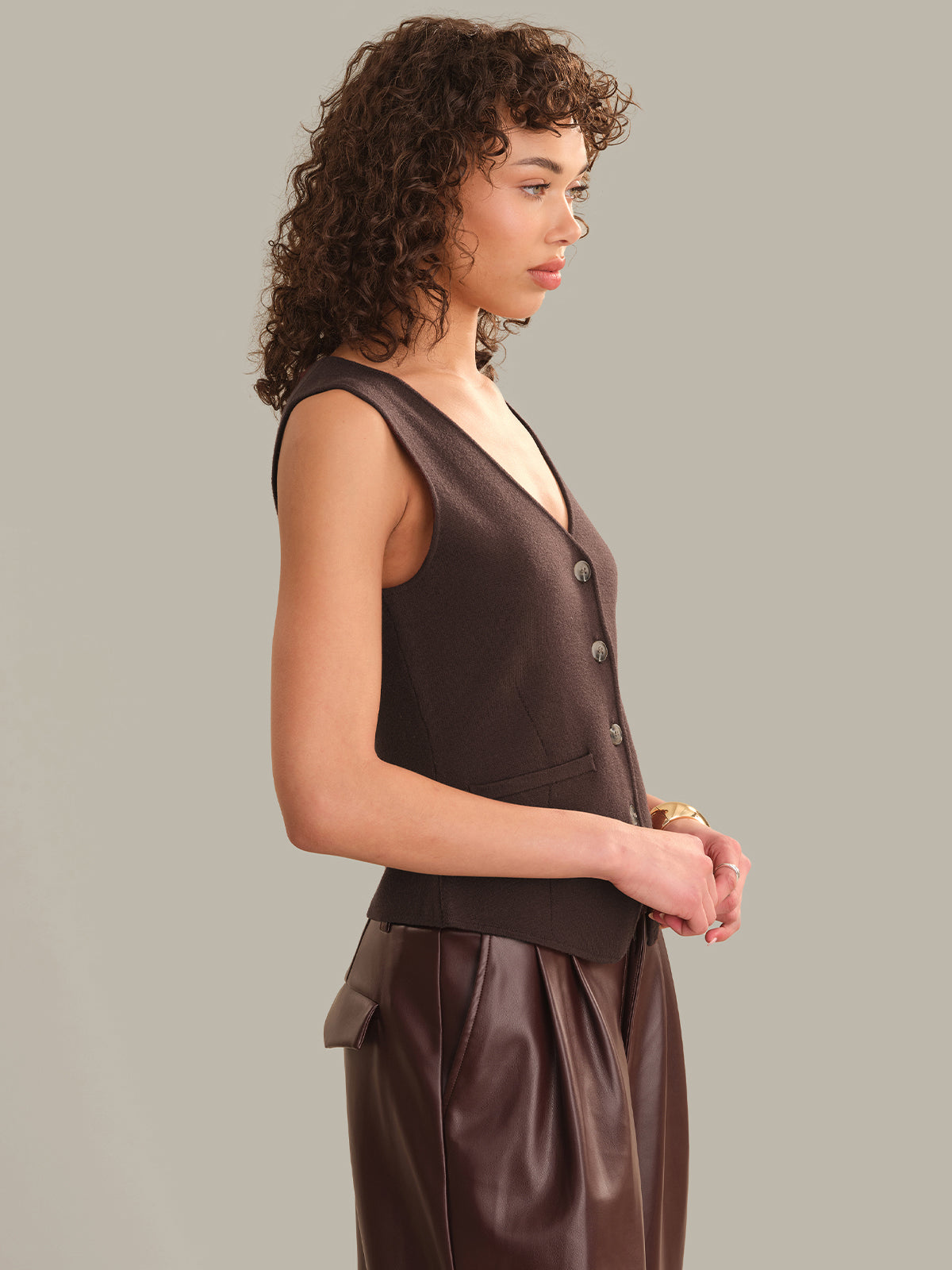 A model with curly brown hair is shown in profile wearing a dark brown sleeveless vest with front buttons and matching dark brown draped bottoms, set against a solid light gray studio background.