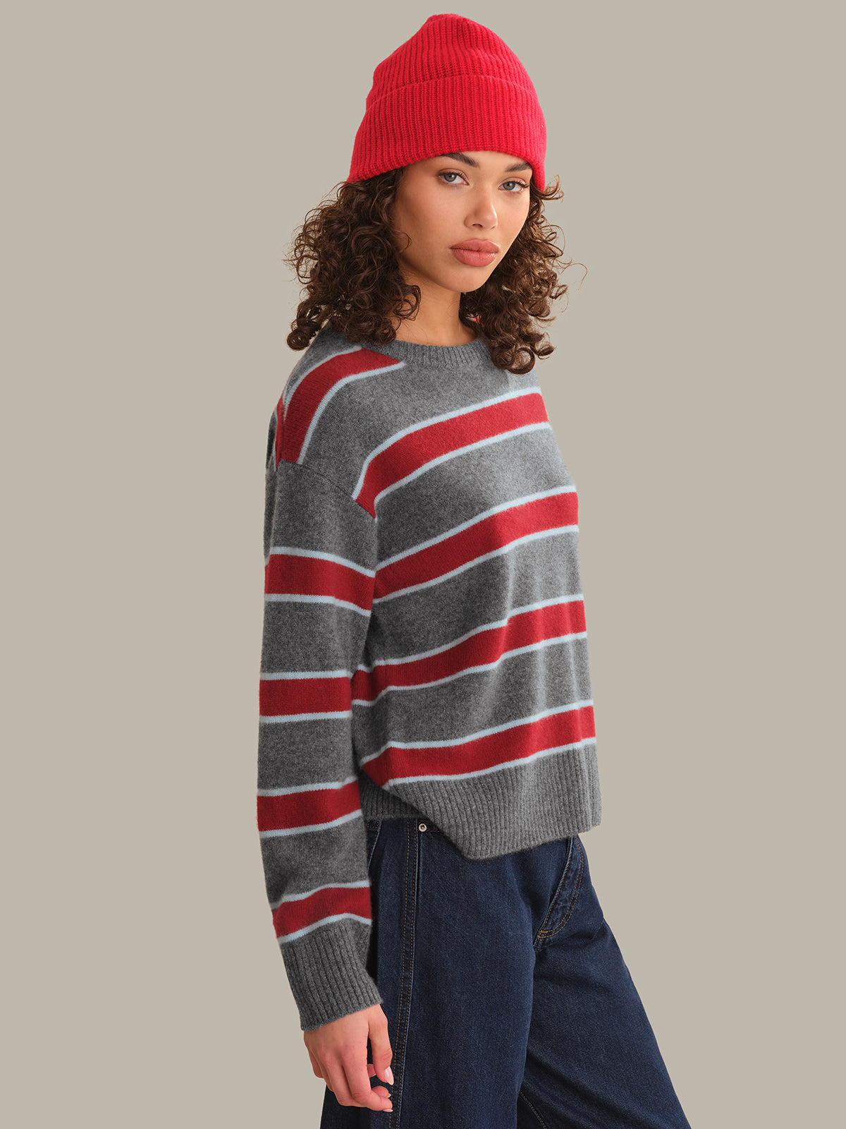 A woman wearing a gray knit sweater with red and white horizontal stripes, a bright red beanie, and dark blue jeans, photographed against a solid light gray studio background.