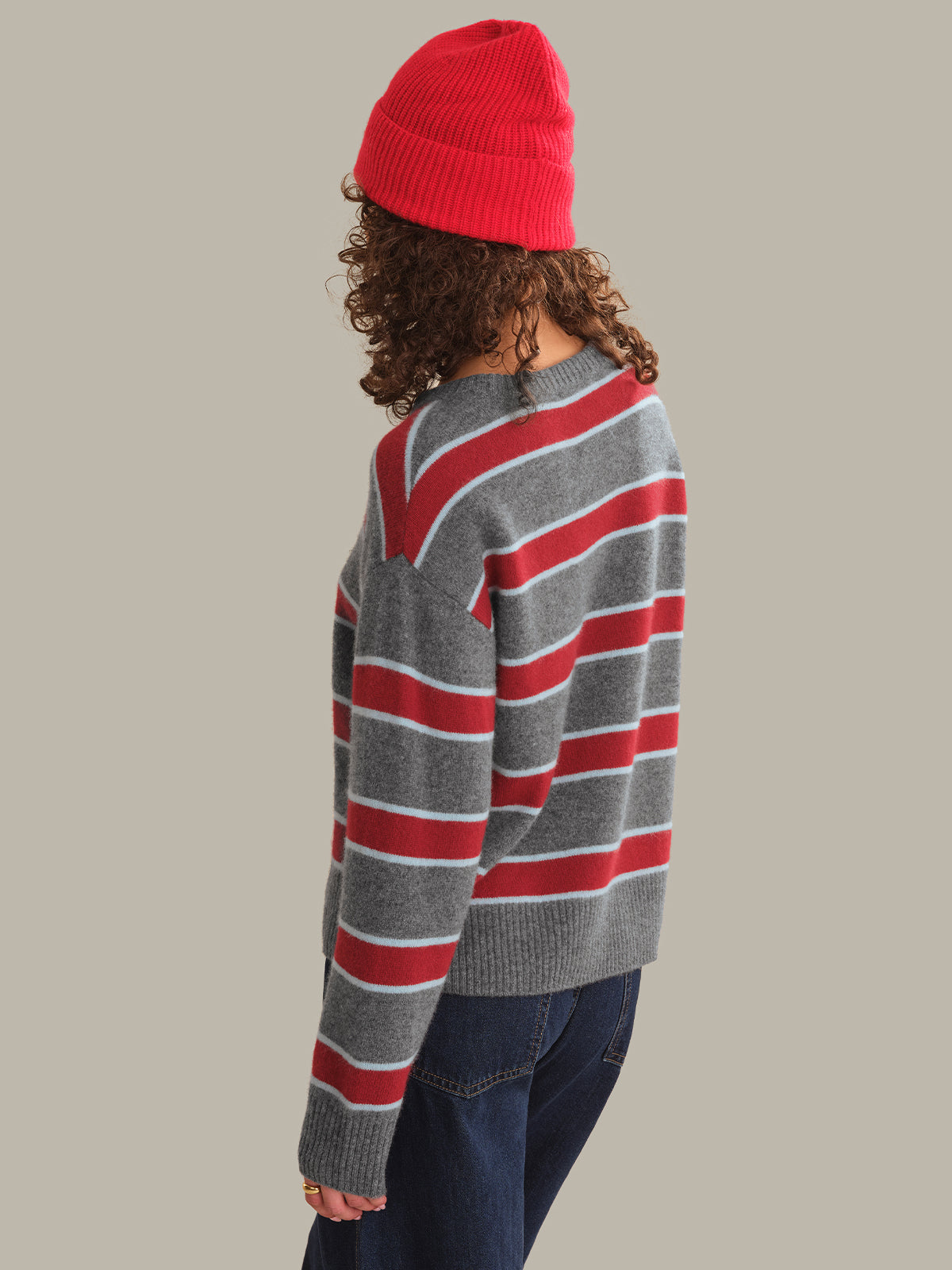 A person shown from the back wearing a gray knit sweater with bold red and white horizontal stripes, a bright red beanie, and dark blue jeans against a solid light gray background.