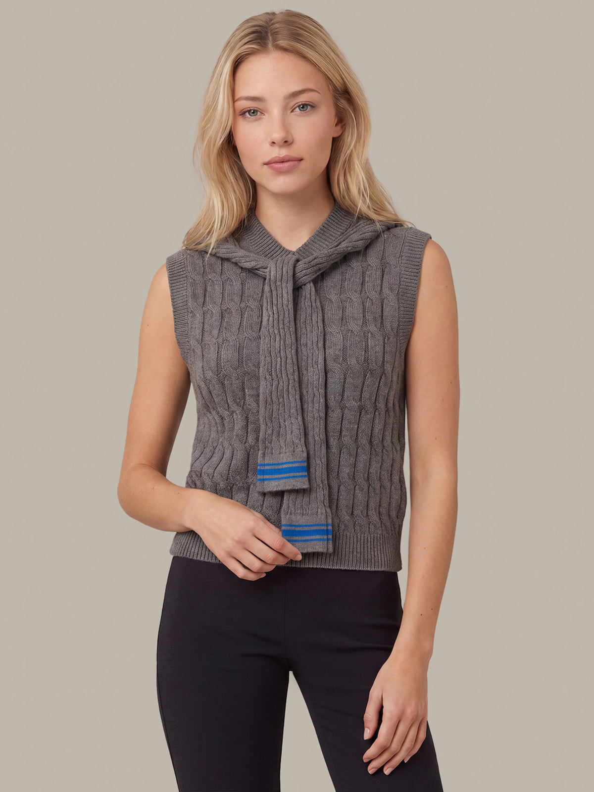 A blonde woman modeling a sleeveless gray cable-knit hooded sweater vest with a matching knotted tie detail and blue striped accents, paired with black pants, against a solid light gray studio background.