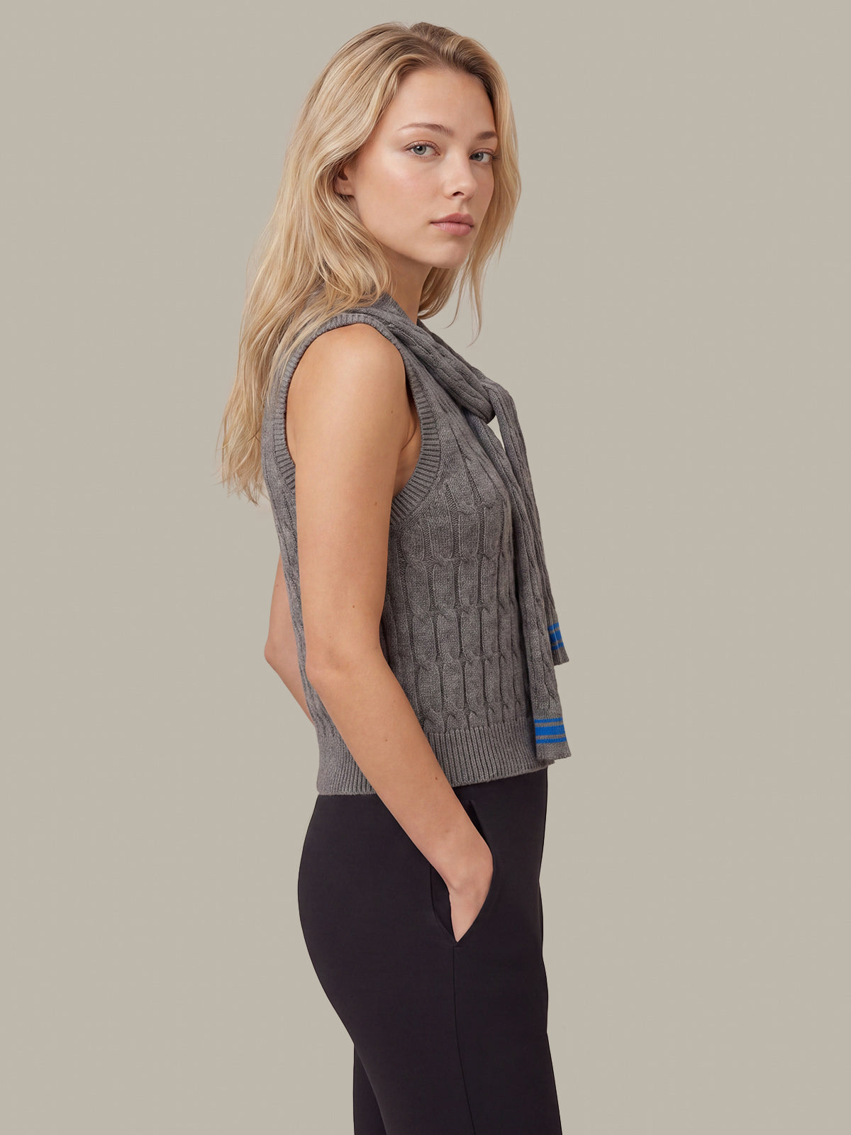 A blonde woman shown in side profile wearing a gray sleeveless cable-knit vest with a matching knit scarf draped over her shoulders, paired with black leggings, against a solid warm gray studio background.