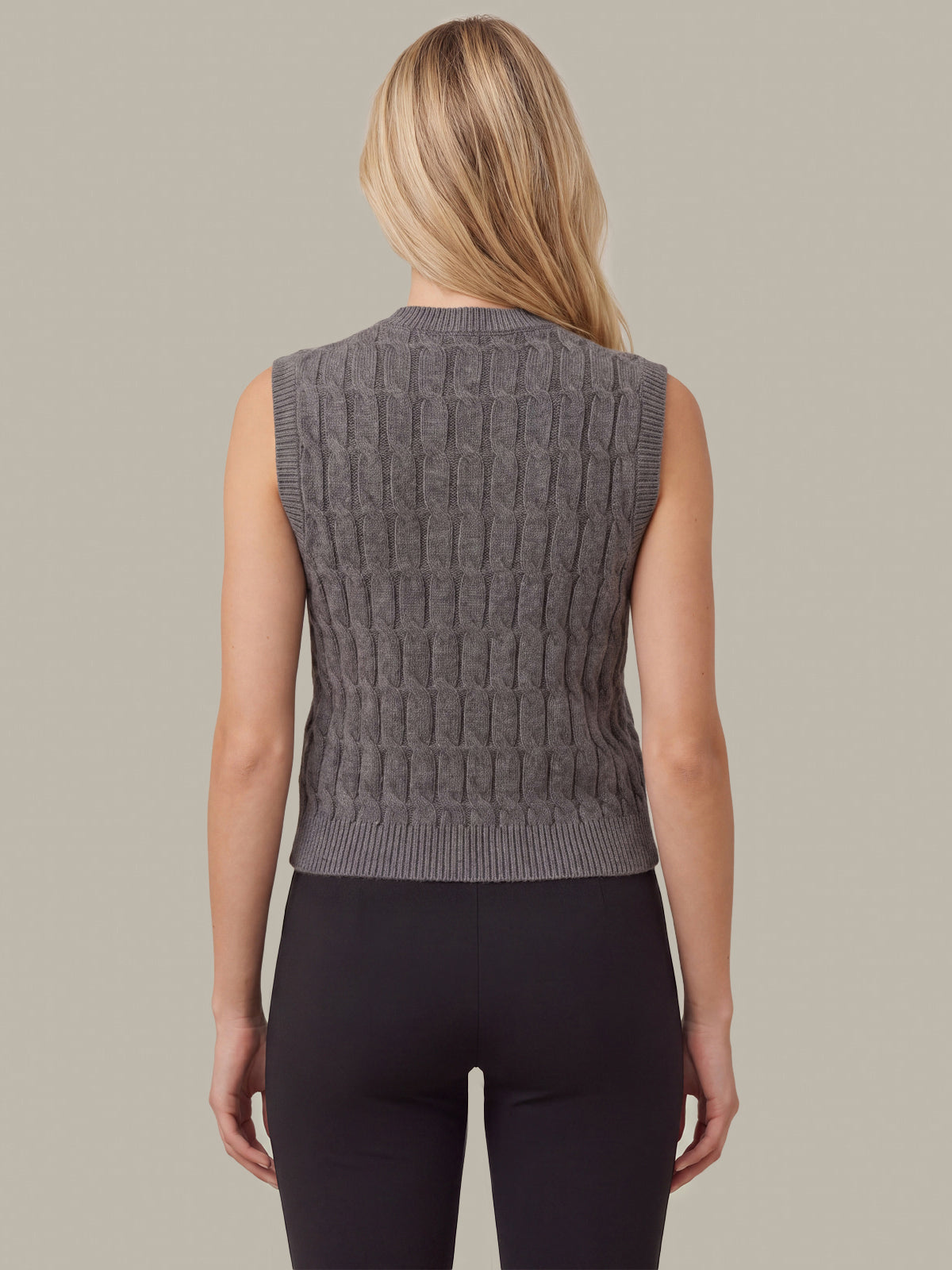 A woman viewed from the back wearing a sleeveless gray cable-knit vest and black leggings against a solid beige background.
