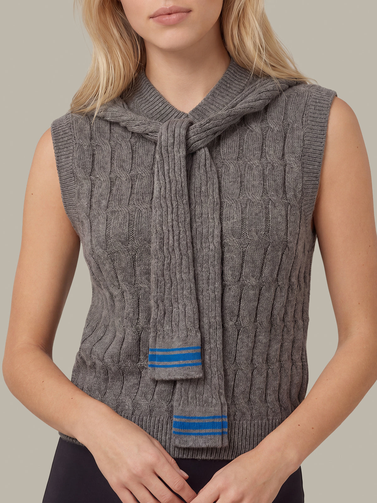 A woman wearing a sleeveless gray cable-knit sweater vest with a knotted tie neckline and blue striped knit details near the hem, shown against a plain beige studio background.