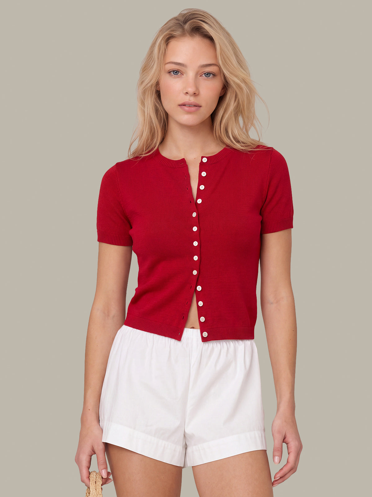 A woman modeling a short-sleeve red knit top with a split neckline and a front button closure, paired with white shorts, against a solid light gray background.