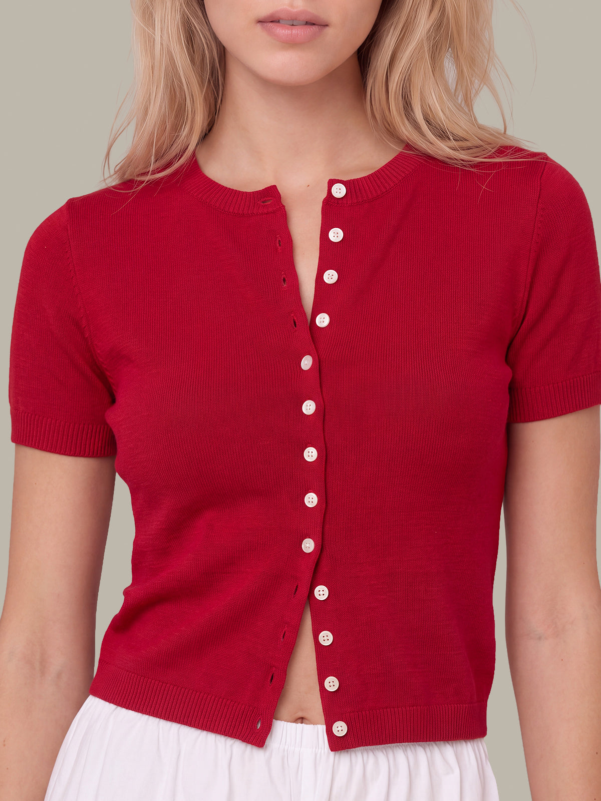 A woman wearing a short-sleeve red knit cardigan top with a buttoned front closure, paired with a white skirt, against a neutral beige background.